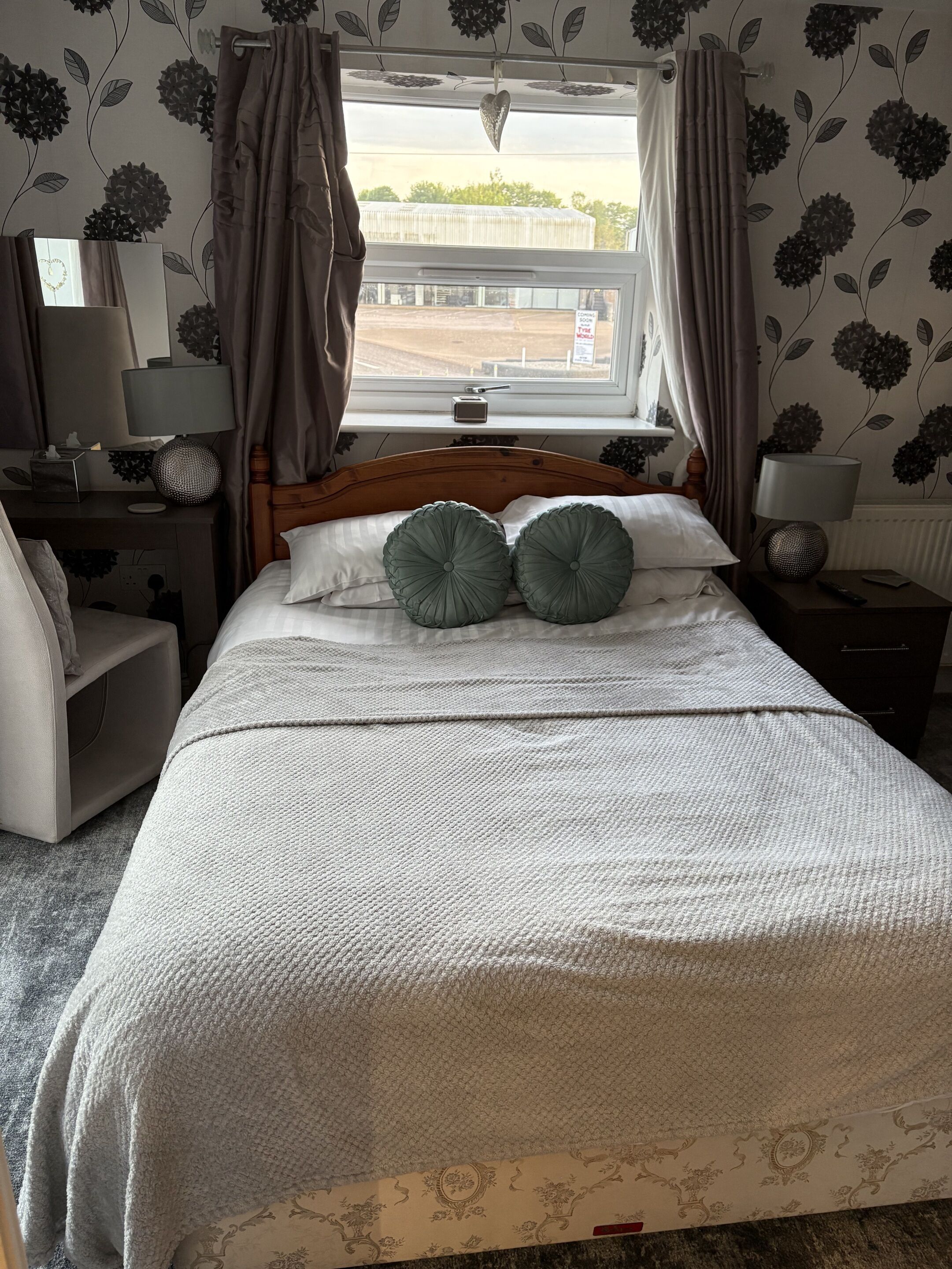 A neatly made bed with two pillows, a bedside table with a lamp, a nightstand with a lamp, and a window with curtains.