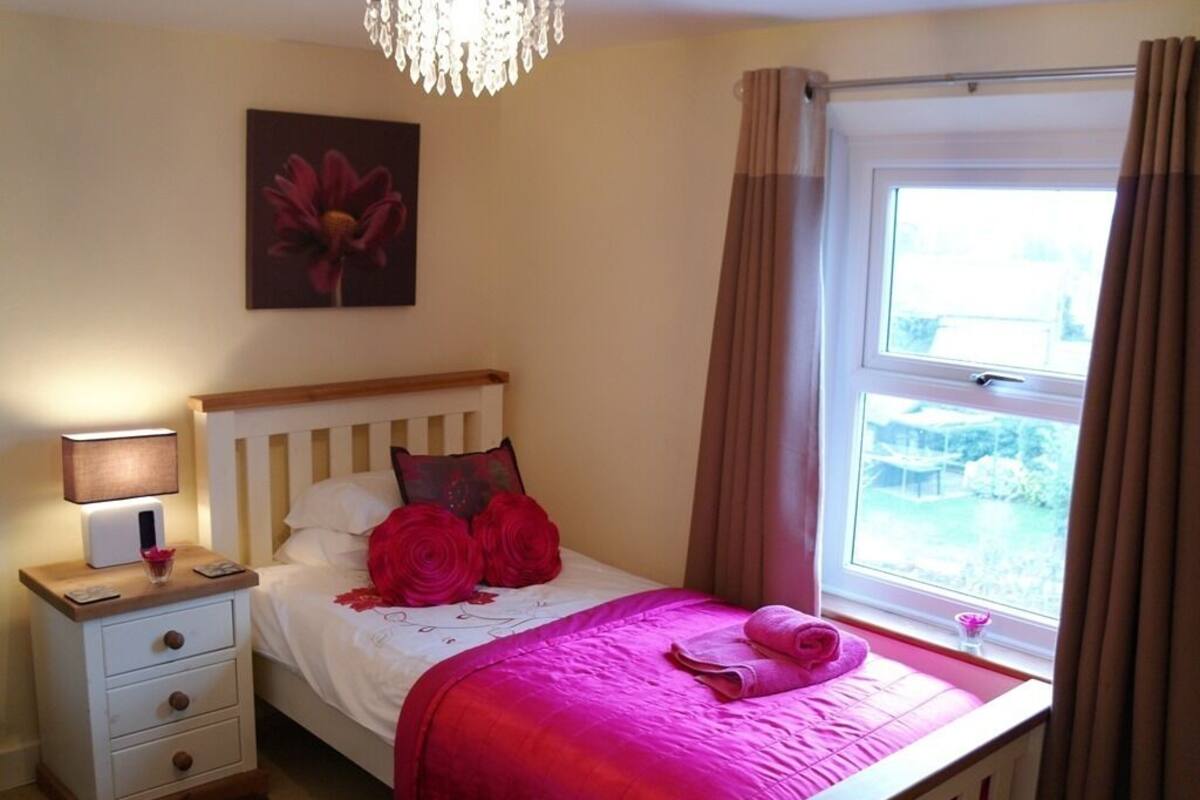 Um quarto com cama, mesa de cabeceira, abajur, janela com cortinas e um quadro na parede.