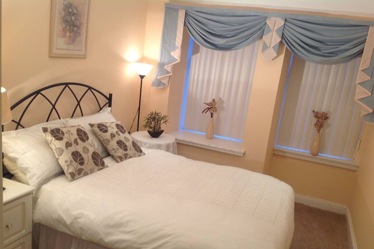 Una cama bien hecha con ropa blanca y almohadas estampadas, una mesita de noche con lámpara, y una ventana con cortinas.