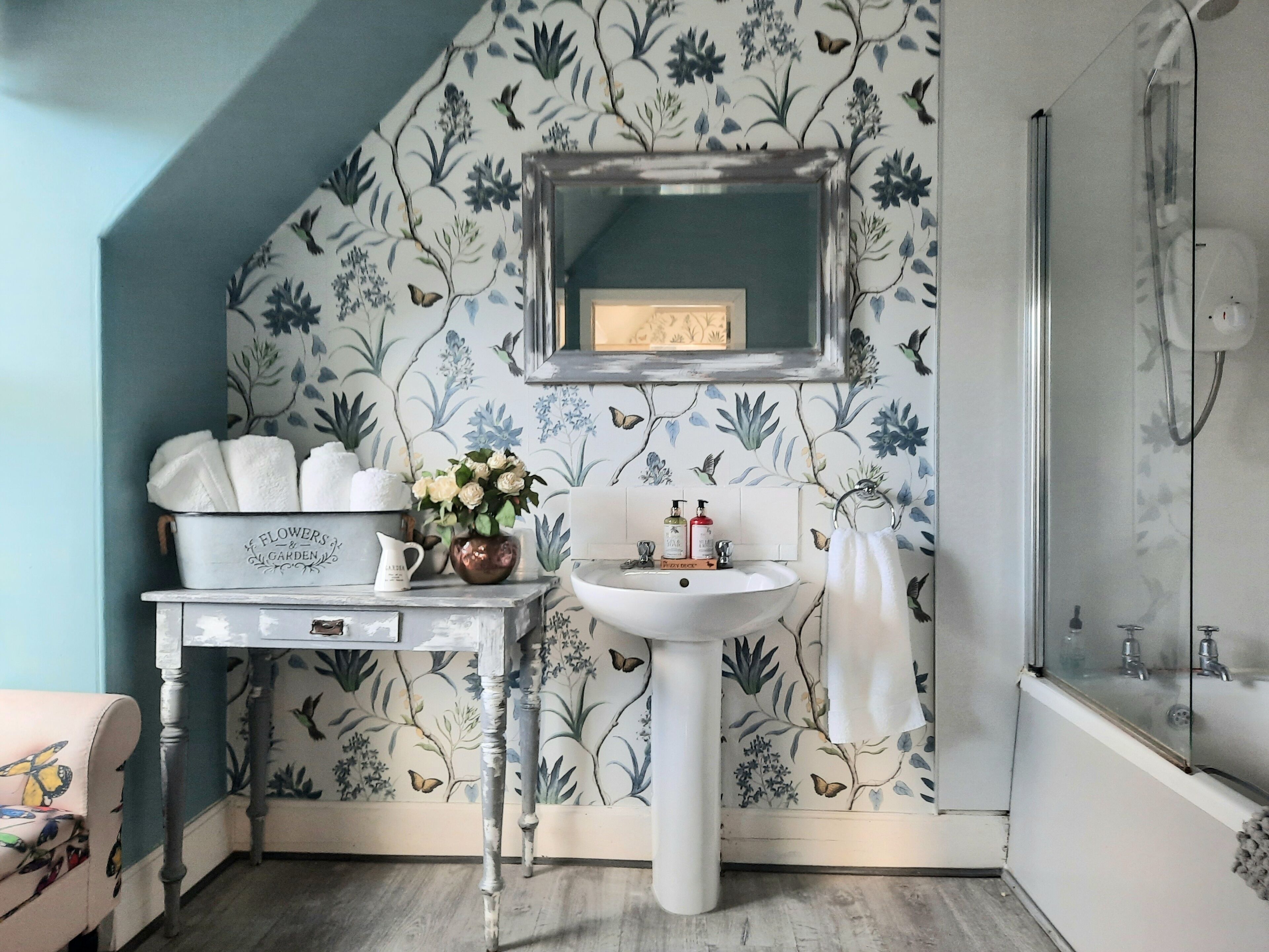 Un baño con papel tapiz floral, un lavabo blanco, un espejo sobre el lavabo y una zona de ducha.