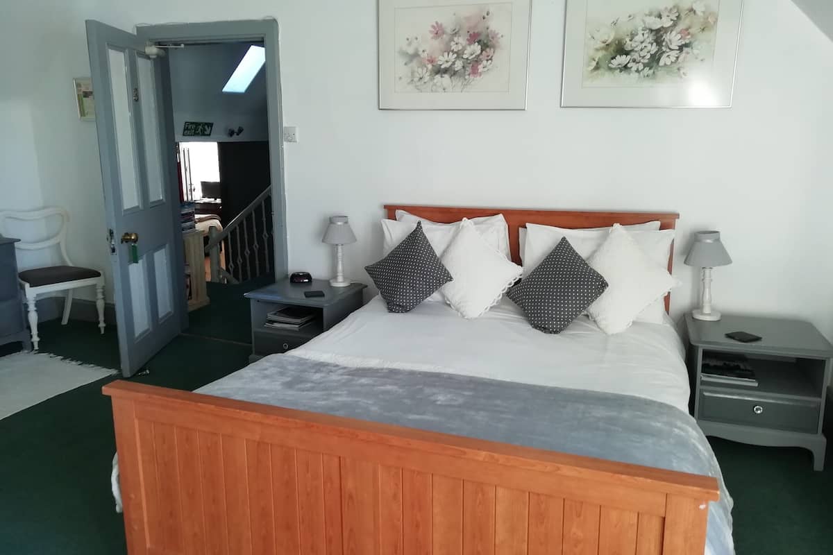Quarto com cama de madeira, duas mesas de cabeceira, um criado-mudo, uma cadeira e porta que dá para outro cômodo.