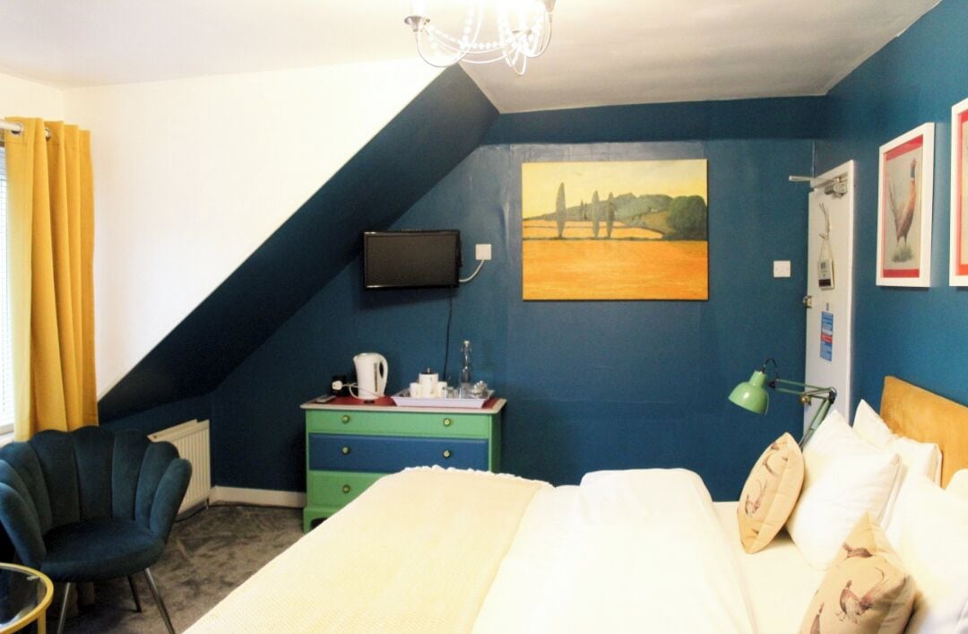 A bedroom with a sloped ceiling, a bed with white bedding, a green dresser, a chair, a painting, and a television on the wall.