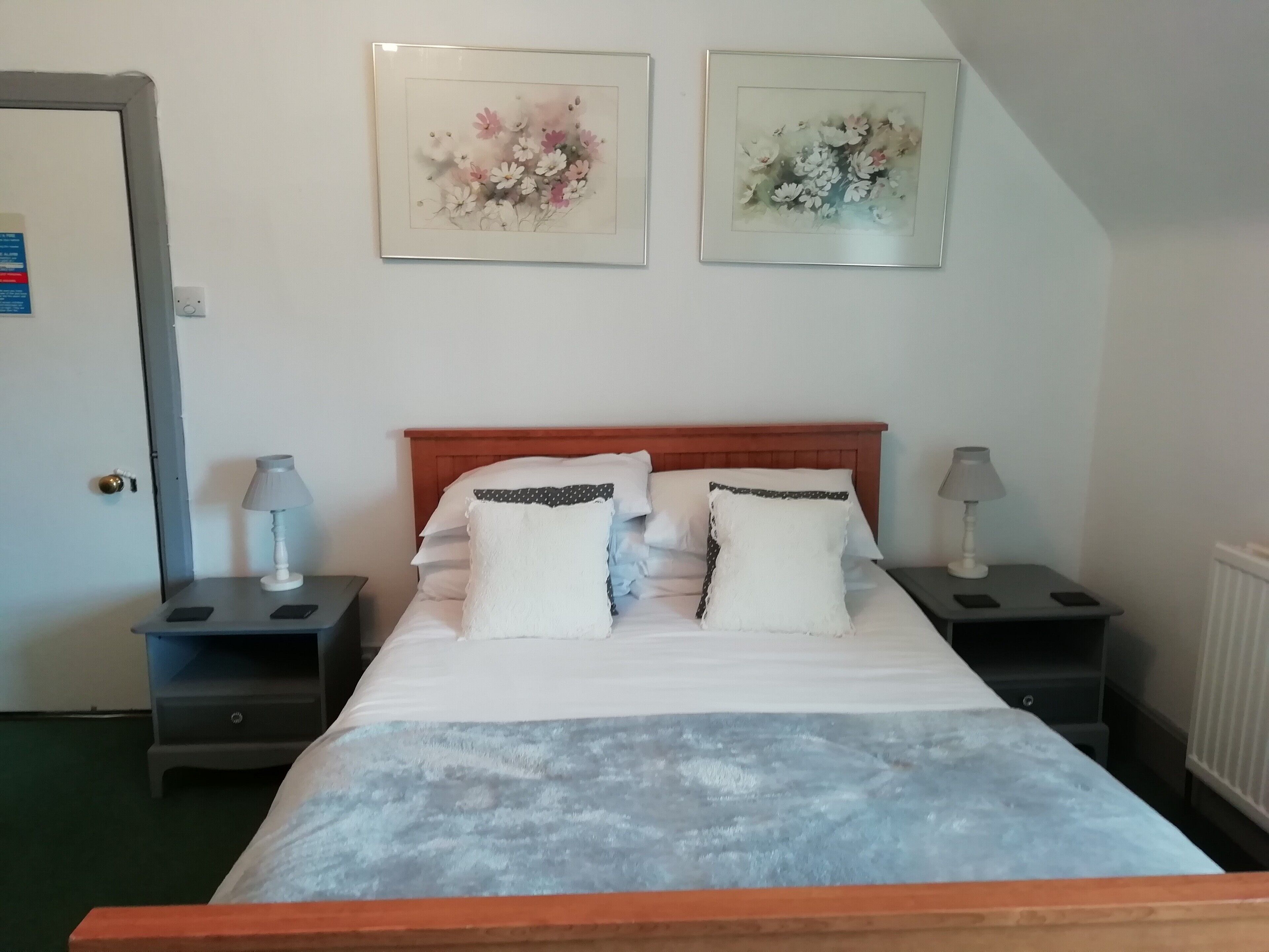 Cama bem arrumada com roupa de cama branca e duas almofadas, com mesas de cabeceira laterais com abajures, em um quarto com cabeceira de madeira e duas obras de arte floral emolduradas na parede.