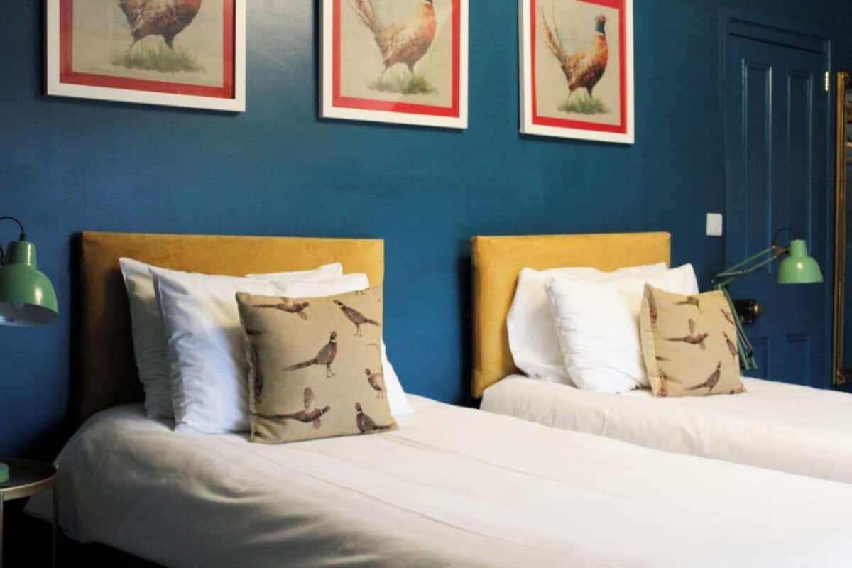 Quarto de hotel com duas camas, cada uma com cabeceira de madeira e roupa de cama branca. Há quadros com galinhas na parede acima das camas.