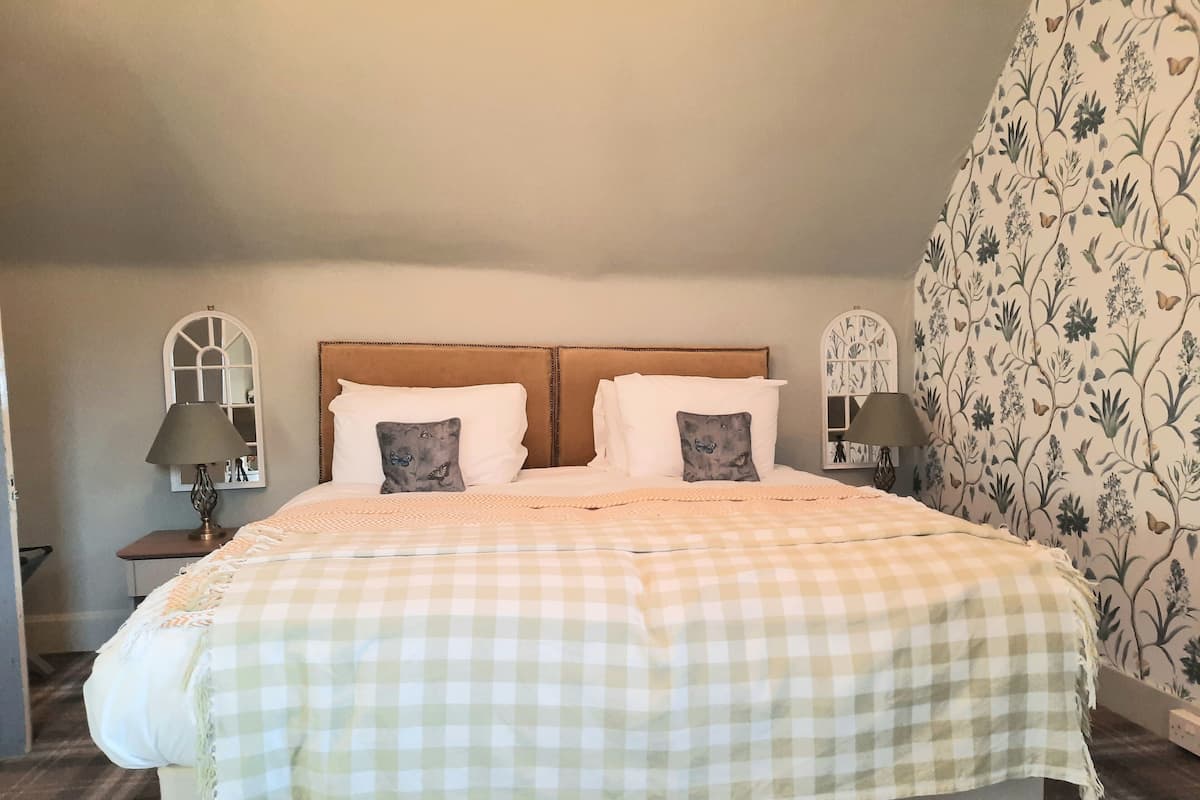 Quarto com colcha xadrez, duas almofadas, cabeceira de madeira e papel de parede floral.