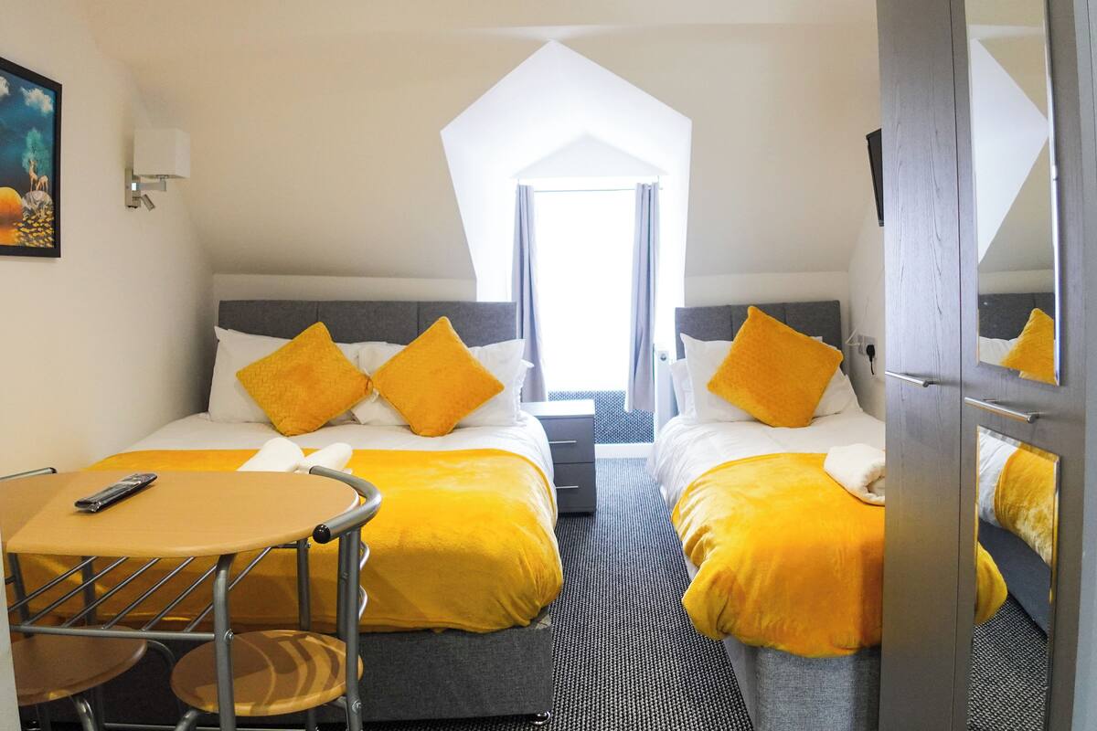 Et hotelværelse med to senge, et lille bord og en stol. Der er et billede på væggen og et vindue med gardiner.