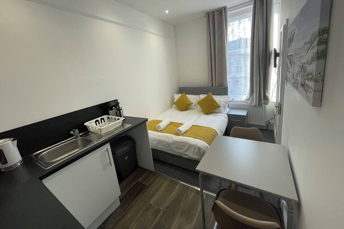 Et kompakt hotelværelse med seng, lille tekøkken, spisebord og et vindue med gardiner.