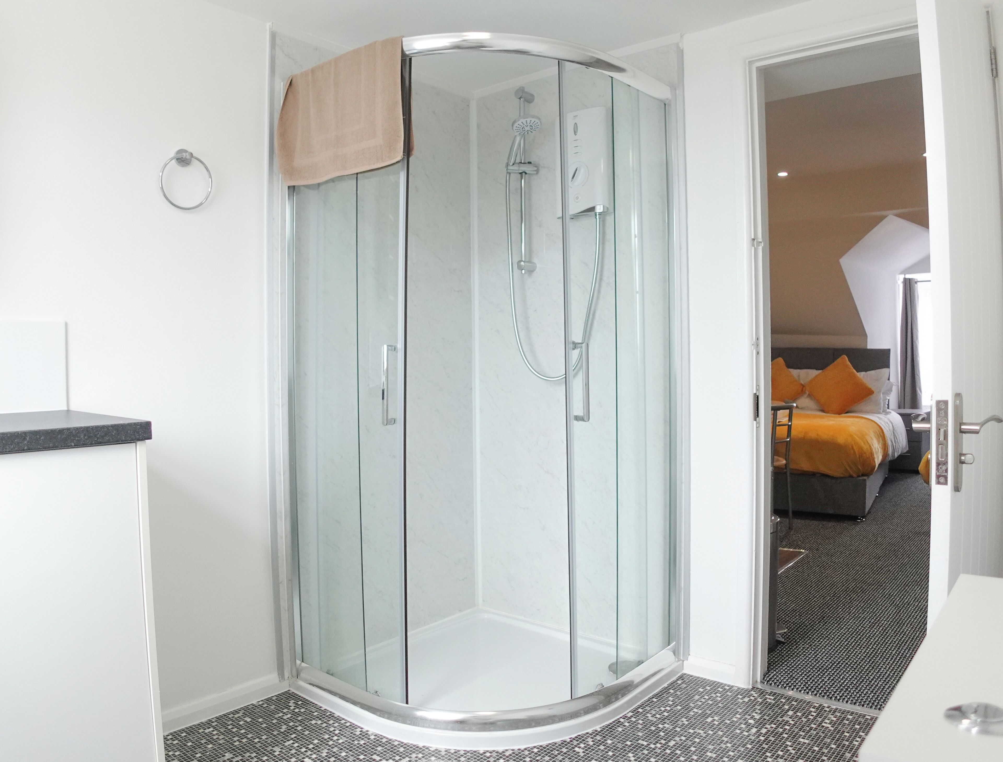 A modern bathroom with a glass-enclosed shower, a towel ring, and a visible bed with orange bedding in the adjacent room.