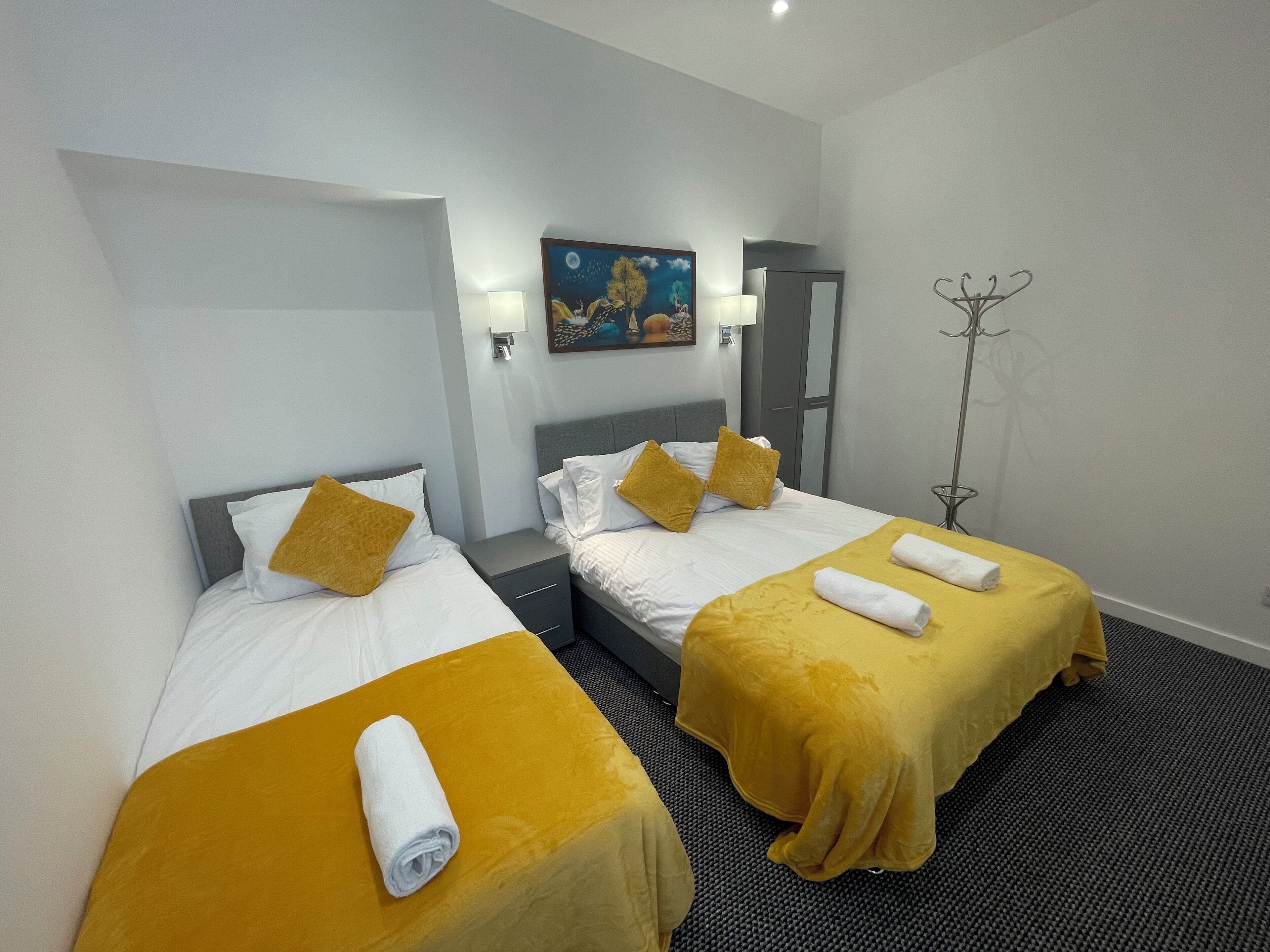Habitación de hotel con dos camas, cada una con cubrecama amarillo y almohadas blancas. Se observa un cuadro en la pared, un armario y una lámpara de pie.
