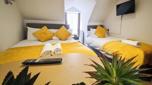 Quarto de hotel com duas camas, cada uma com roupa de cama amarela e almofadas combinando. Há uma televisão na parede, um controle remoto na mesa e um vaso de planta na mesa.