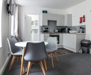 A modern kitchen with white cabinets, a microwave, and a coffee maker. There is a dining area with a round table and four chairs. A fire extinguisher is mounted on the wall. - Station House Plymouth (Plymouth)