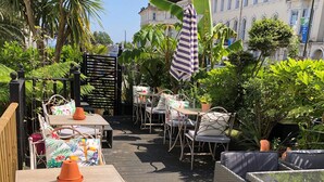 Een terras buiten met tafels en stoelen, omgeven door weelderige begroeiing en een gebouw op de achtergrond.