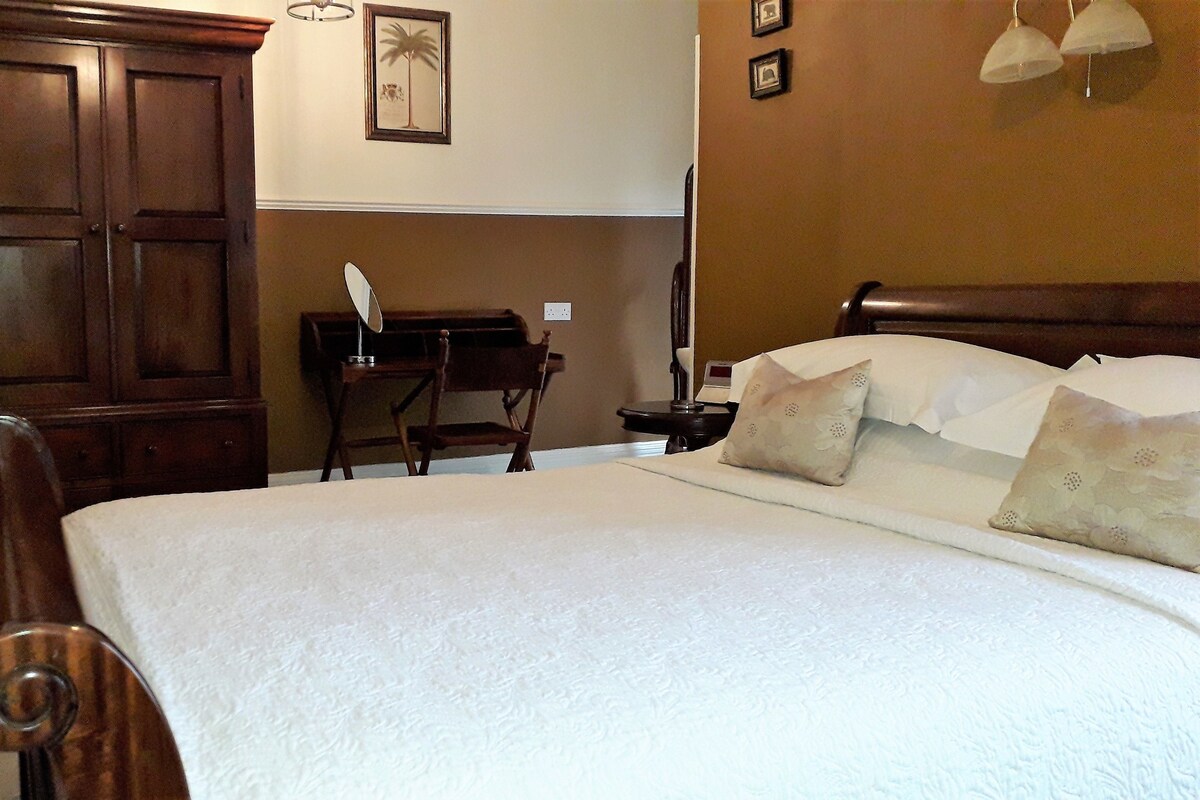 A neatly made bed with pillows, a wooden wardrobe, a desk with a lamp, and a wall clock.