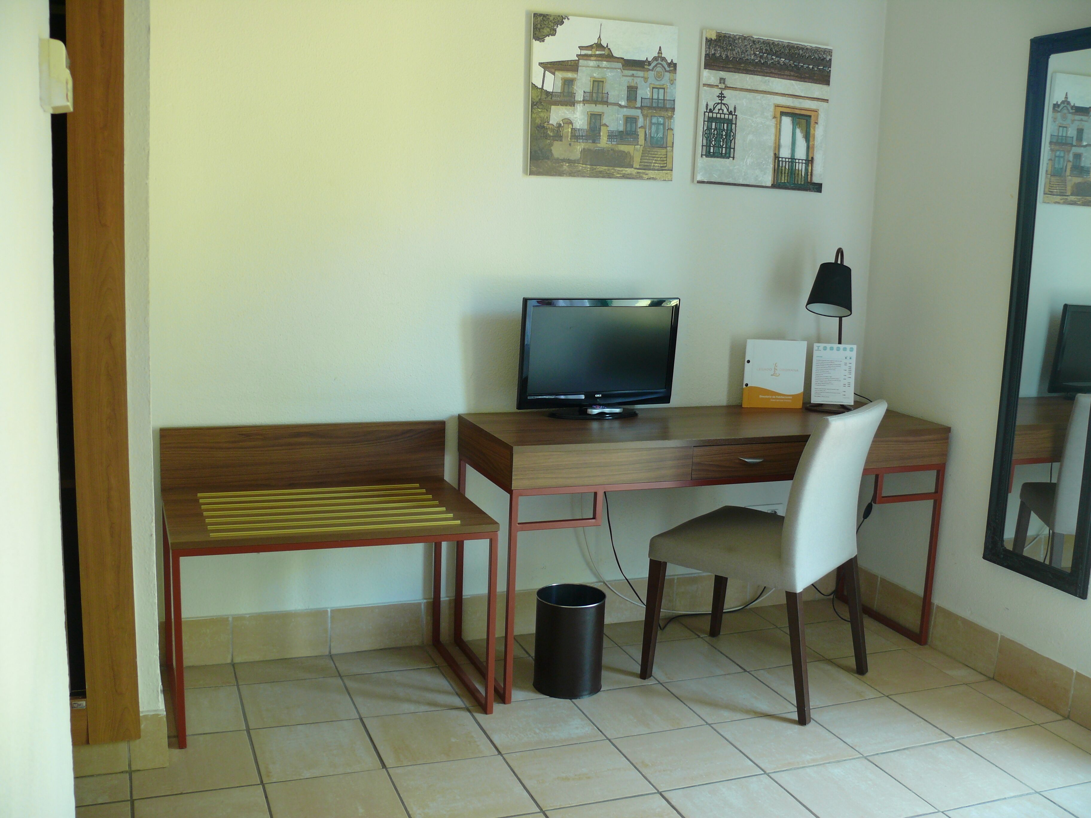 A hotel room with a wooden desk, a chair, a television, a lamp, and a trash can.