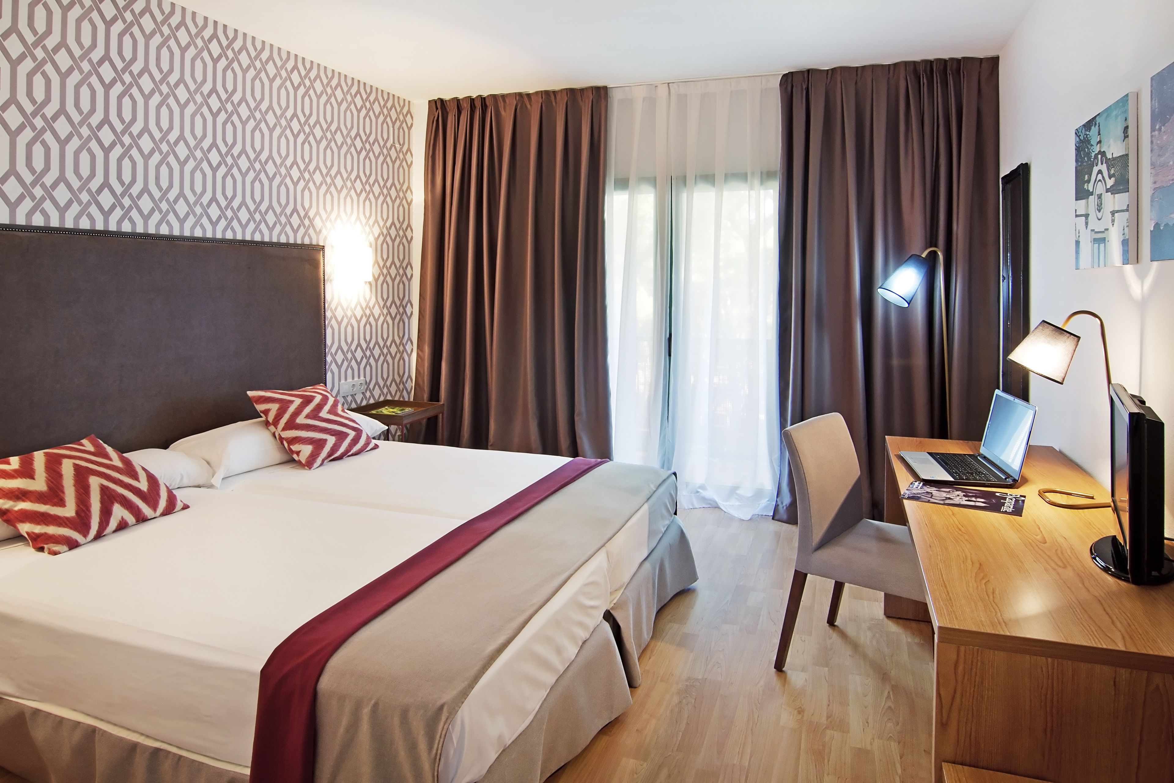 A hotel room with a large bed, a desk with a laptop, a chair, and a desk lamp.