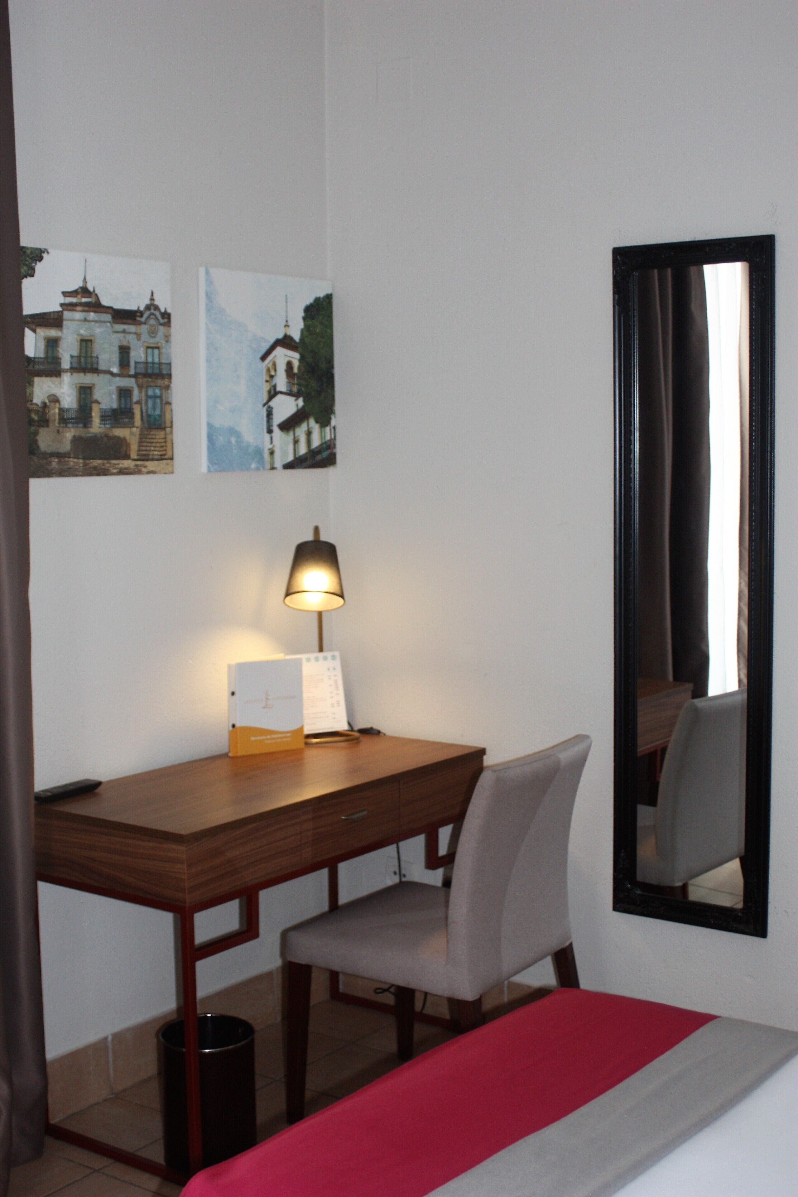 Una habitación de hotel con un escritorio de madera, una silla, una cama con colcha roja y gris, una lámpara de pie y dos cuadros en la pared.