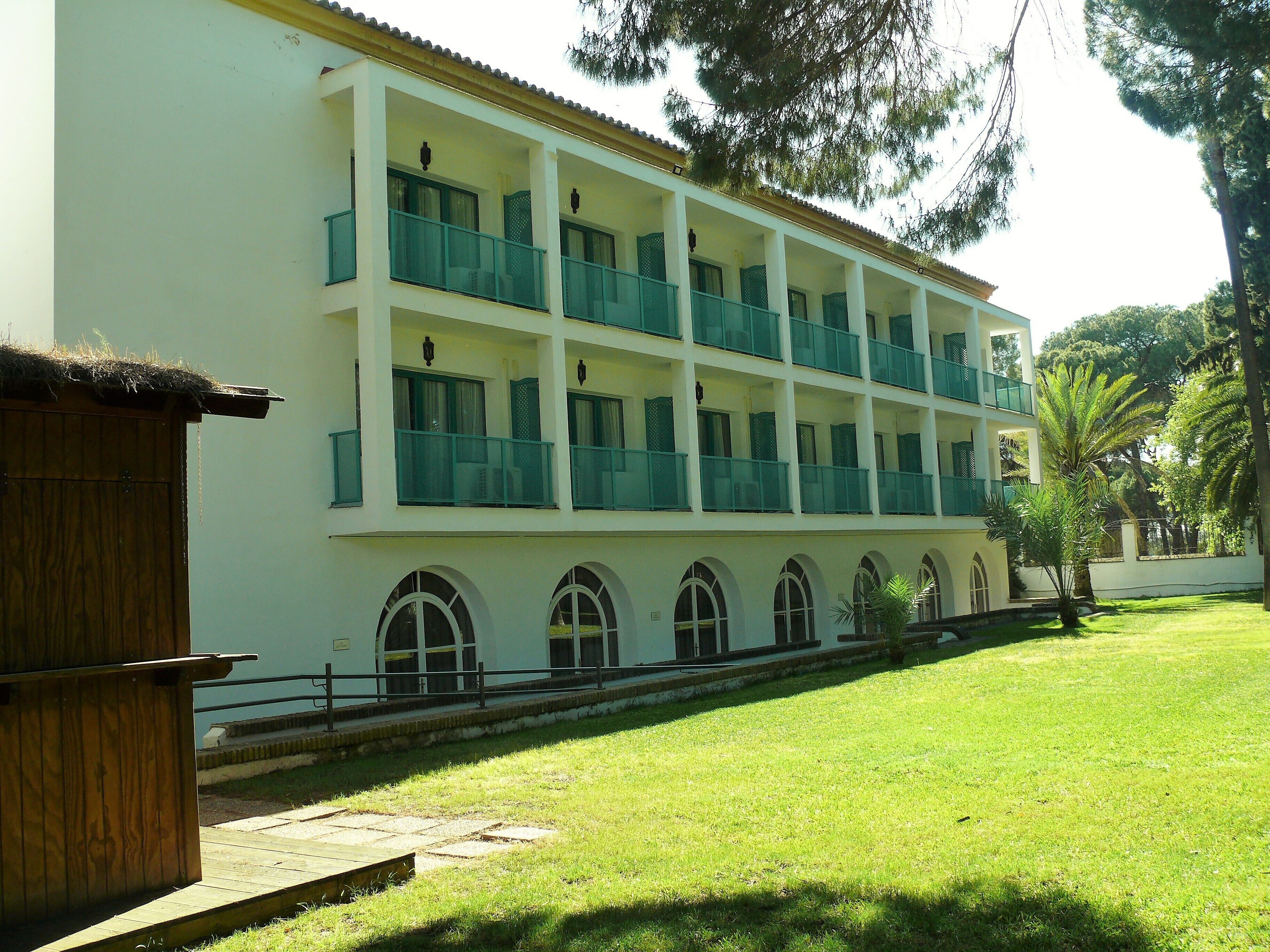 A multi-story building with balconies, a grassy courtyard, and a wooden structure to the left.