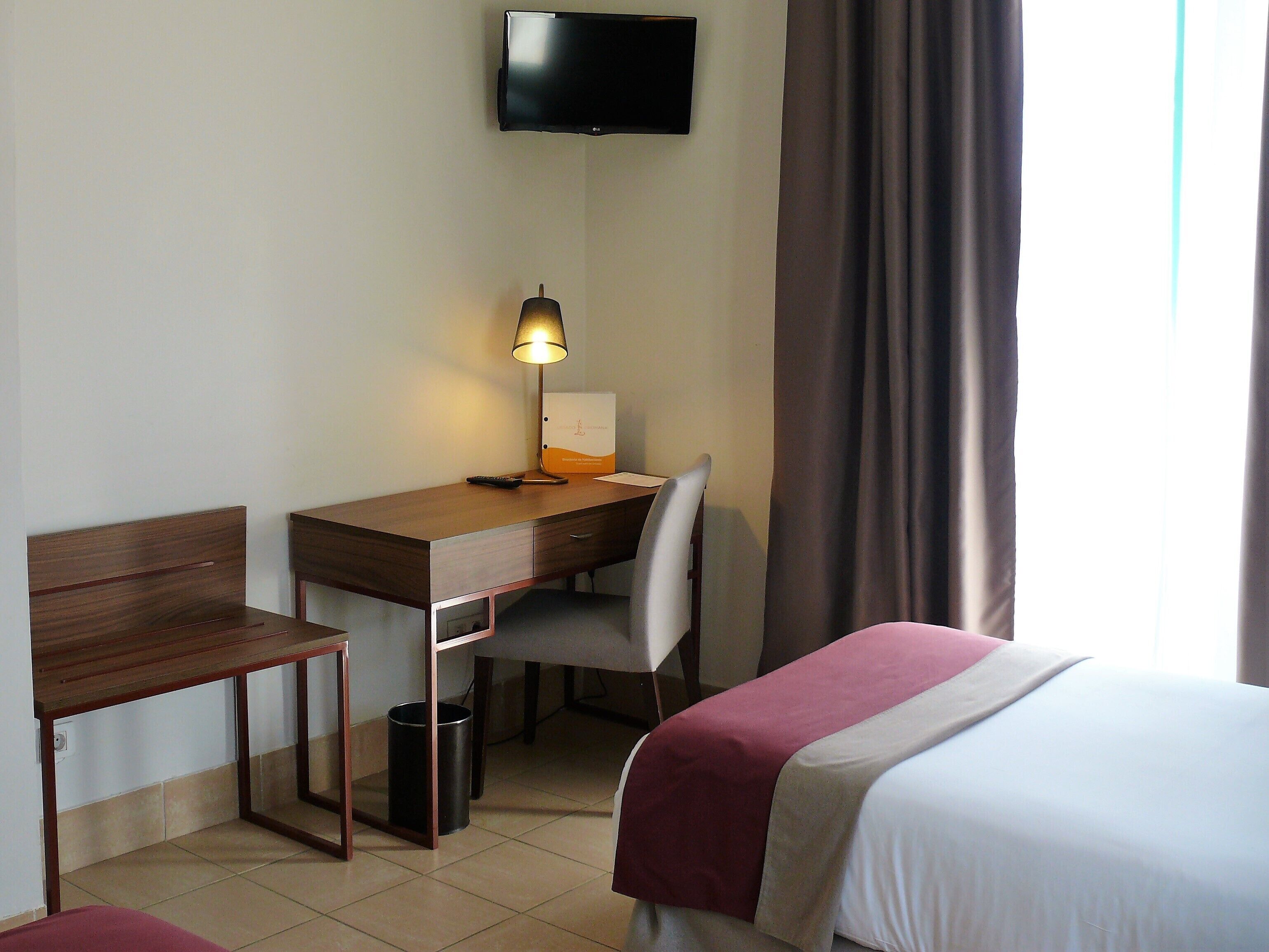 A hotel room with a bed, a desk with a chair, a TV mounted on the wall, and a window with curtains.