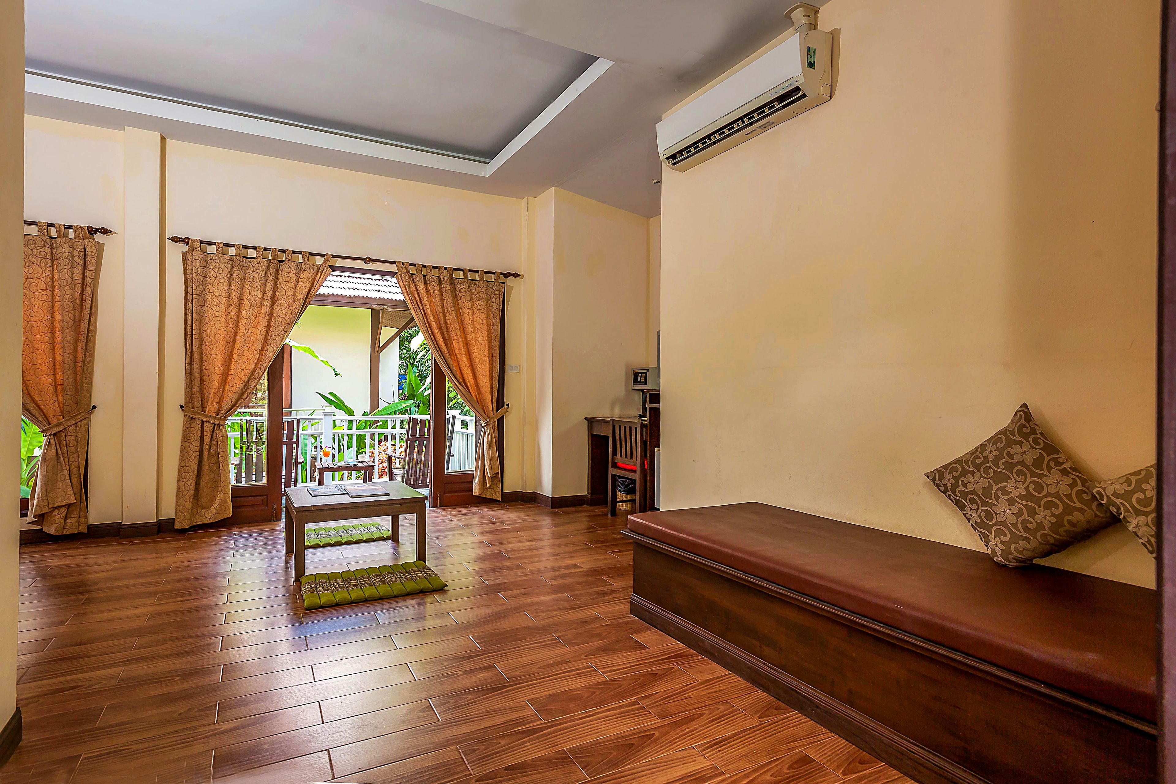 A room with a wooden floor, a bench with a cushion, and a view of greenery outside.