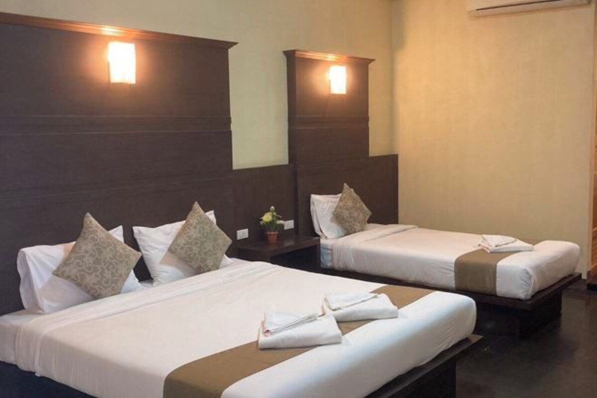 Habitación de hotel con dos camas, cada una con ropa blanca y almohadas color beige. Hay dos mesitas de noche con lámparas, y una pequeña mesa con un florero.