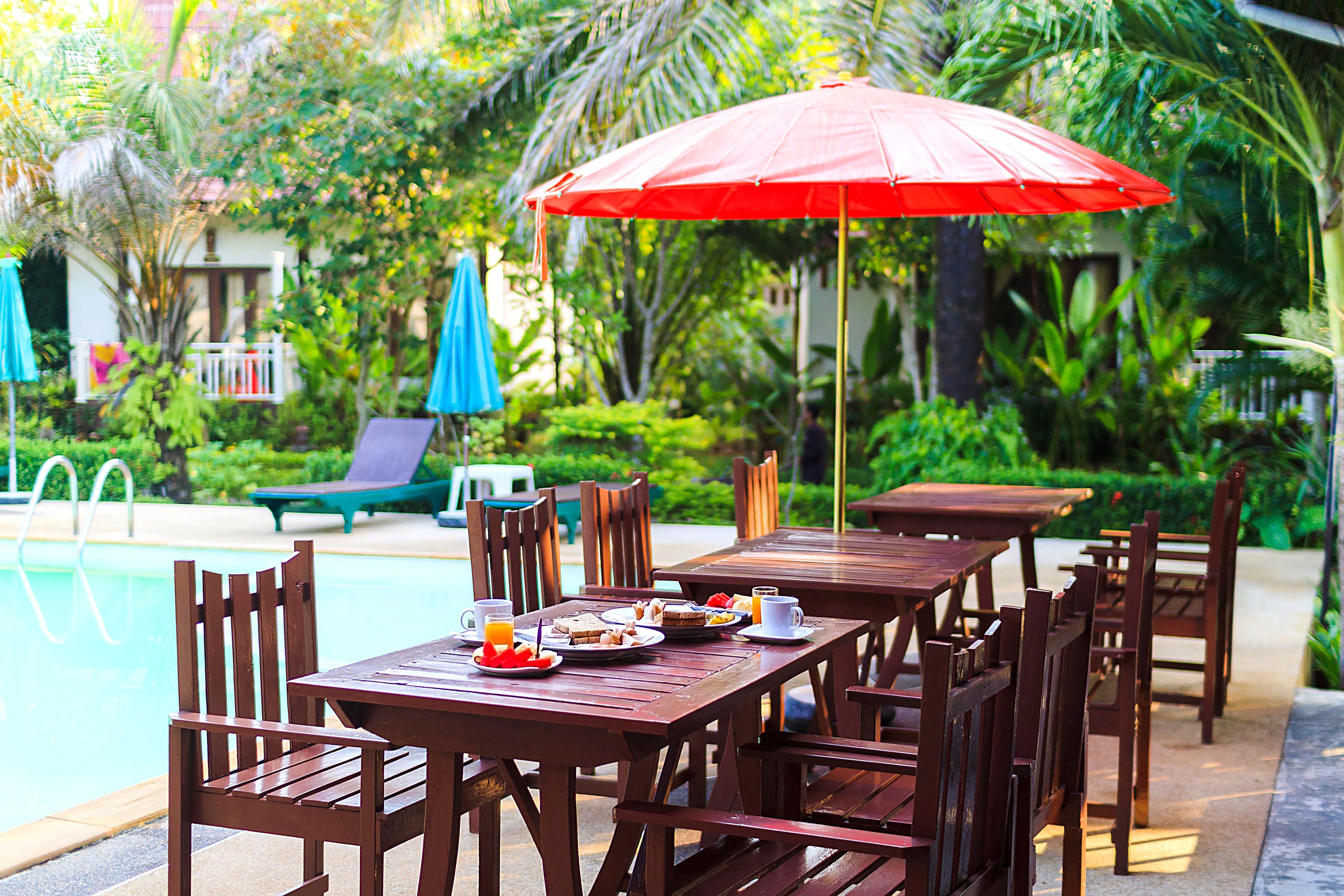 A poolside dining area with wooden tables and chairs, a red umbrella, and a pool in the background.