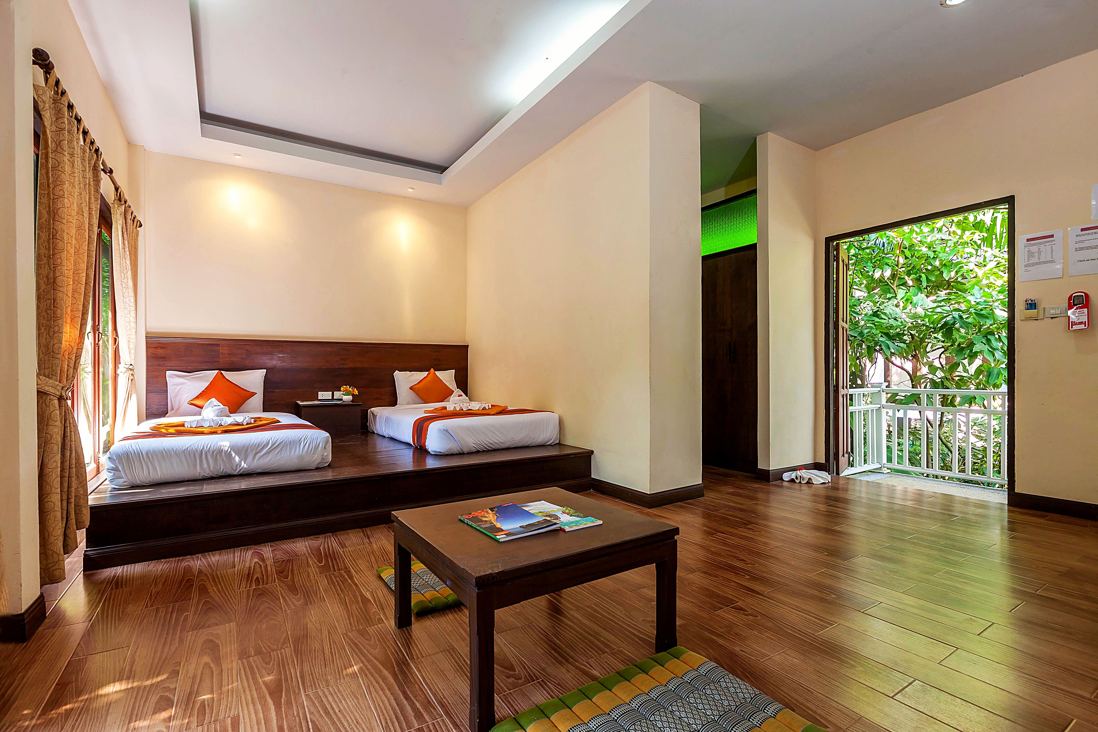 A hotel room with two beds, a wooden floor, a small table, and a view of greenery outside.
