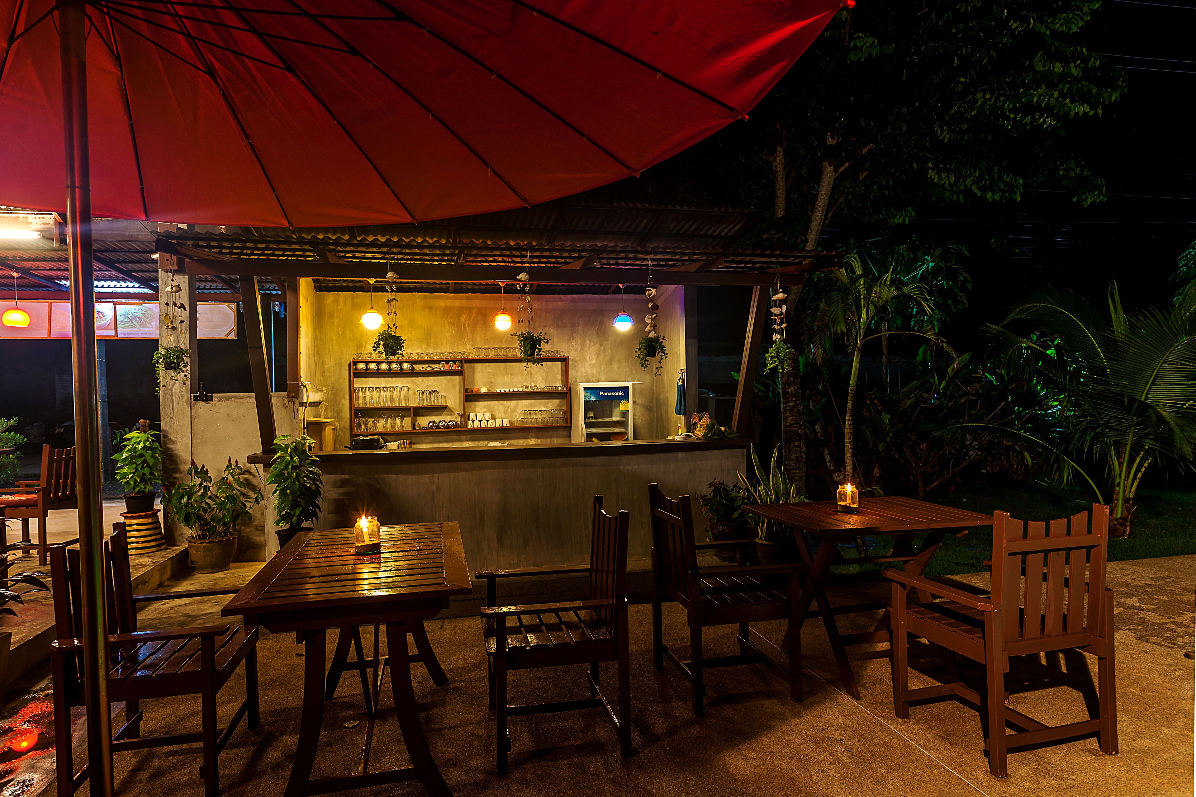 An outdoor seating area with wooden tables and chairs, a red umbrella, and a bar counter with various items and plants.