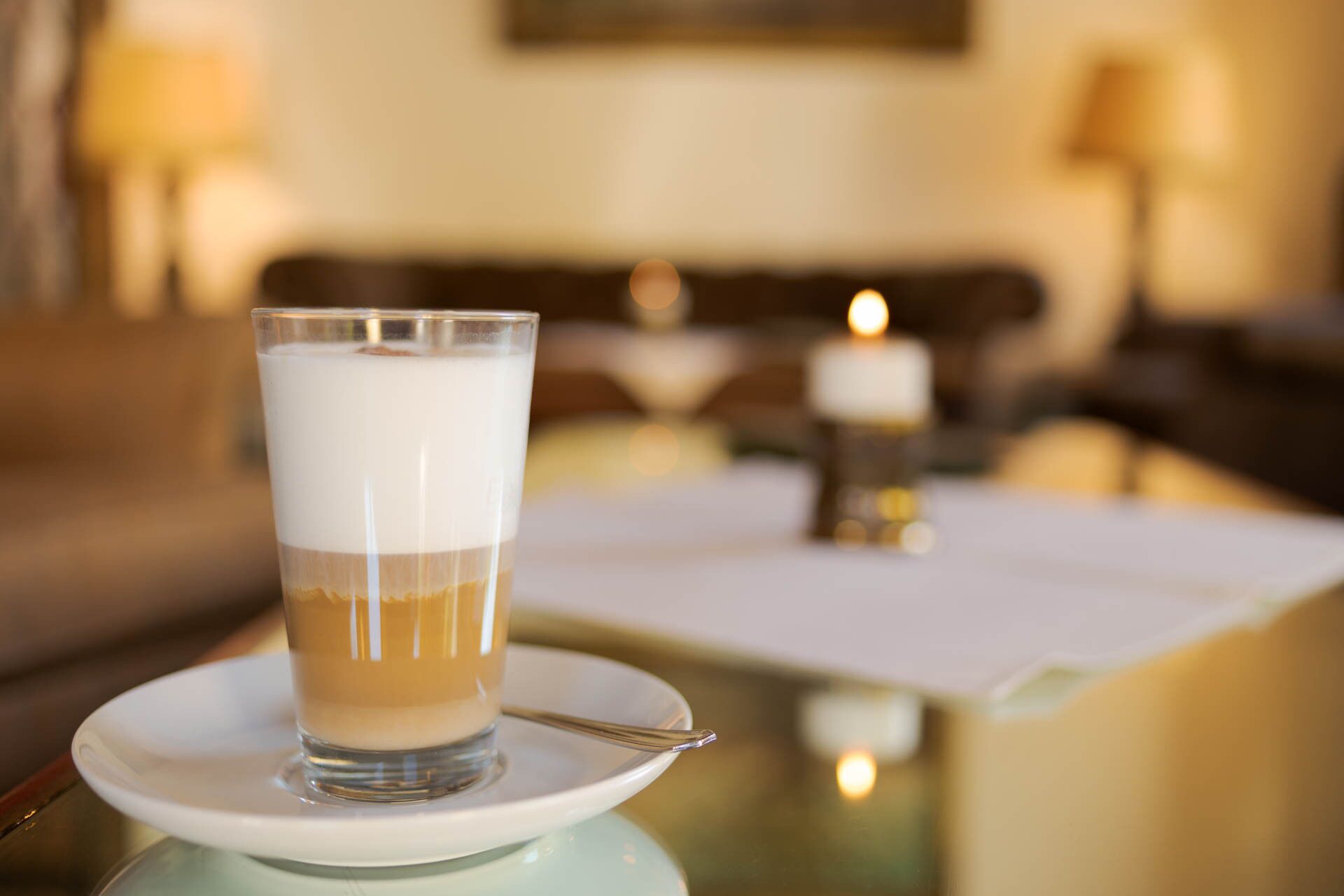 Een glas gelaagde drank op een schotel, met een lepel erop, in een goed verlichte kamer met een tafel en een brandende kaars op de achtergrond.