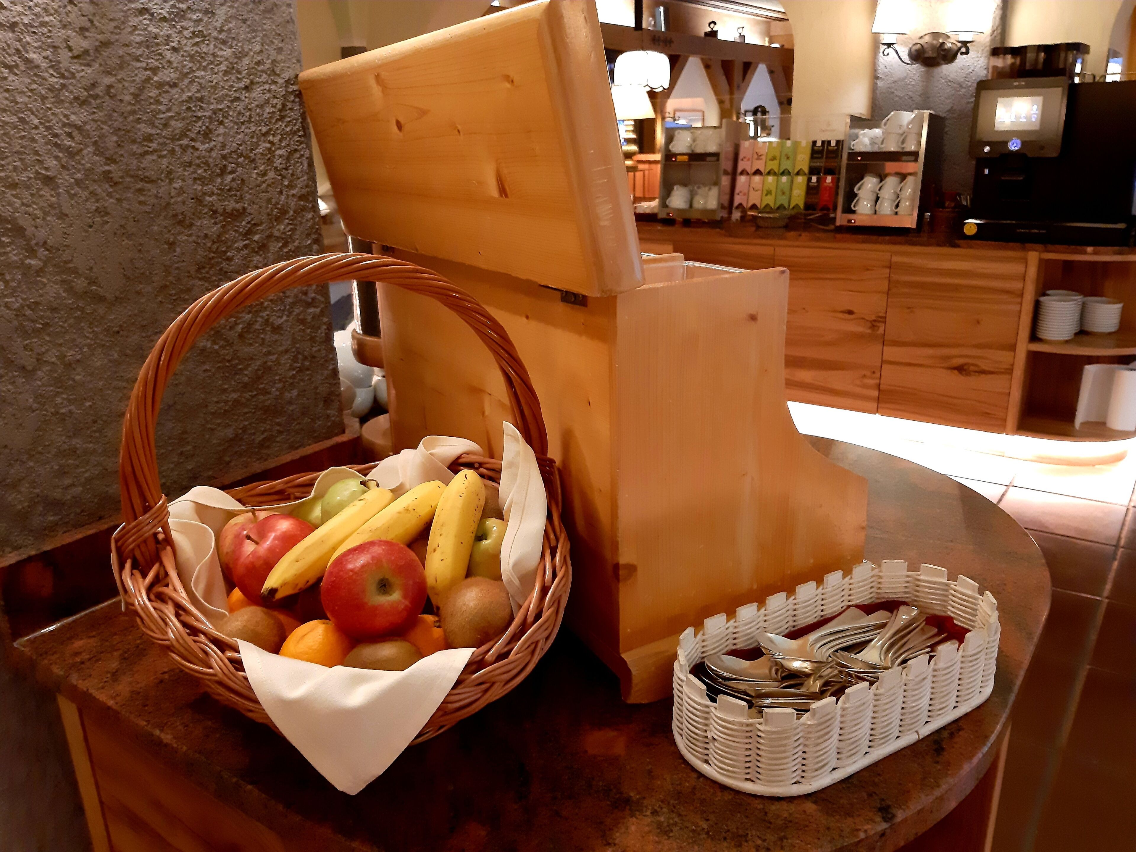 A wicker basket with fruits and a napkin on a table, a white plastic container with utensils, and a coffee machine in the background.