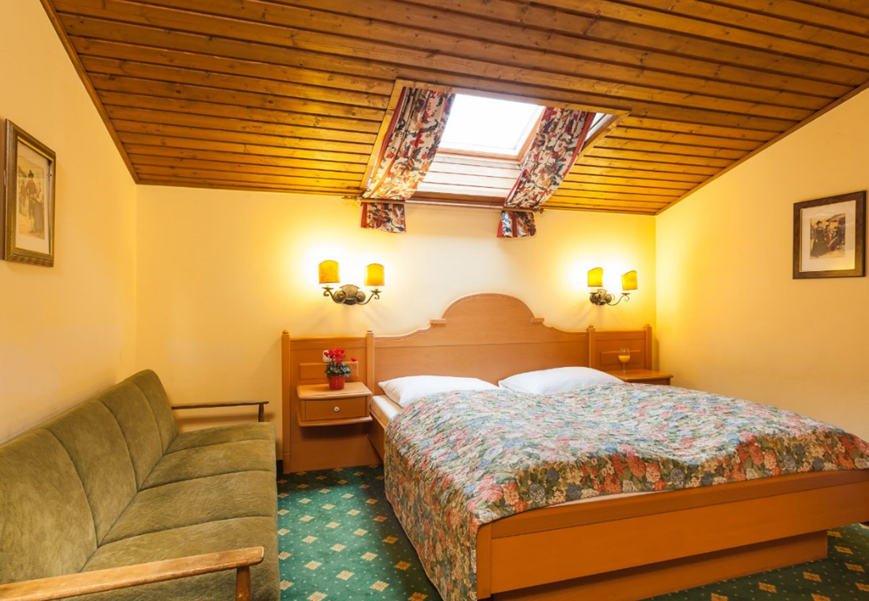 Un dormitorio con techo de madera, una cama con colcha floral, dos lámparas de noche, una mesita de noche con un florero y un sofá verde.
