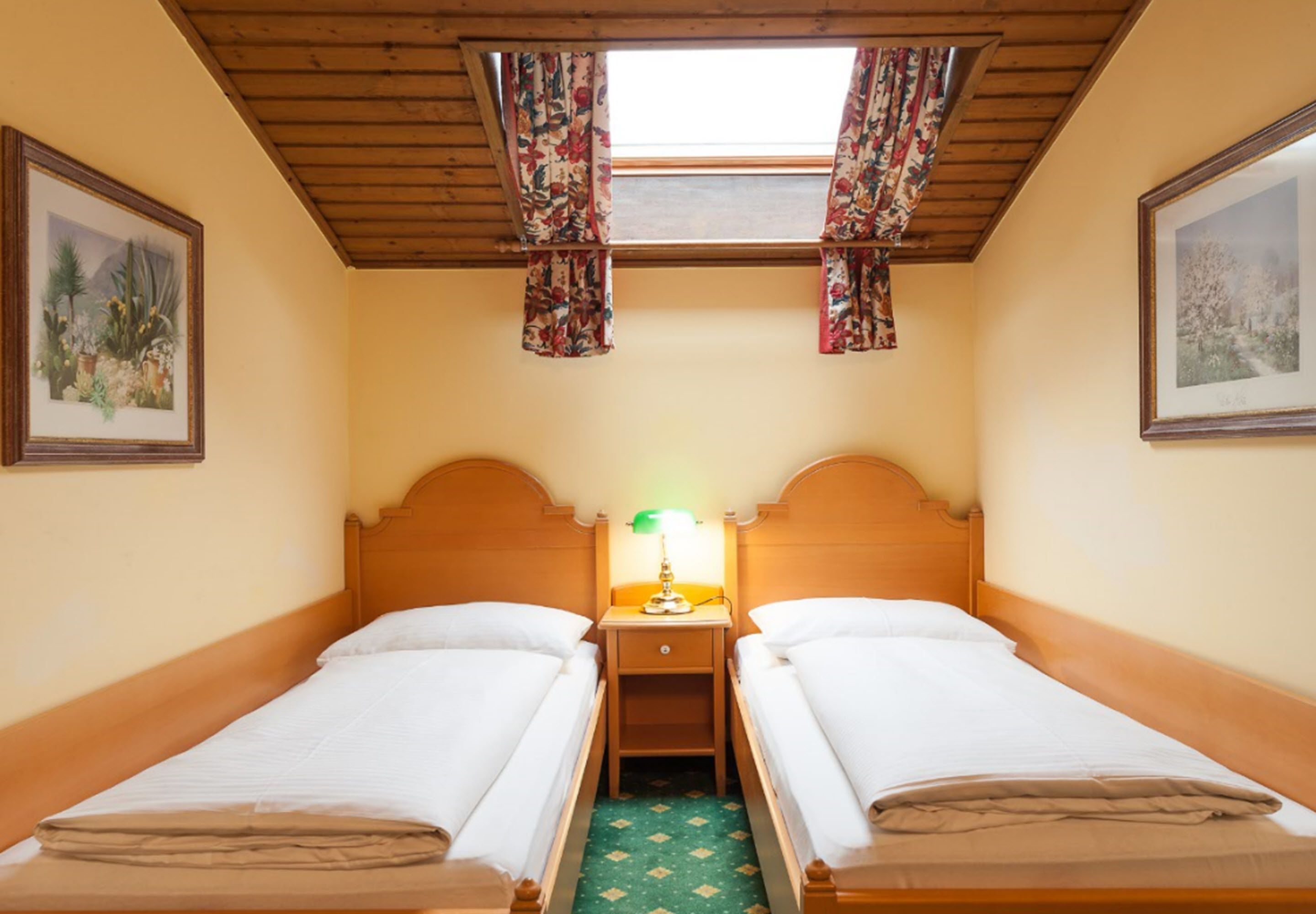 Una habitación con dos camas individuales, una mesita de noche con lámpara, una ventana con cortinas de motivos florales y cuadros en las paredes.