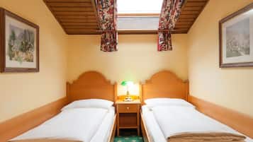 Una habitación con dos camas individuales, una mesita de noche con lámpara, una ventana con cortinas de motivos florales y cuadros en las paredes.