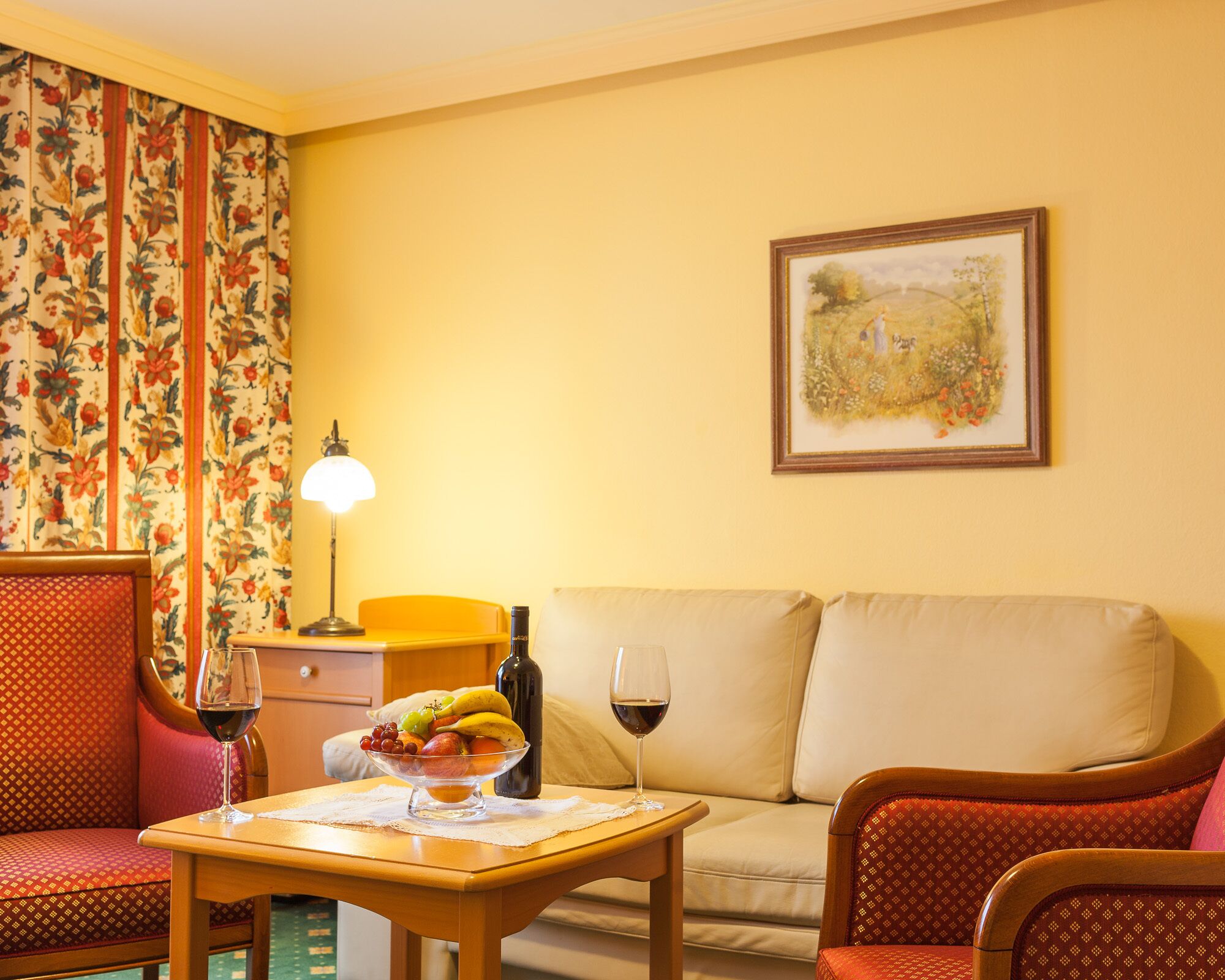 A cozy living room with a sofa, chairs, a table with a fruit bowl and wine glasses, a lamp, and a framed painting on the wall.