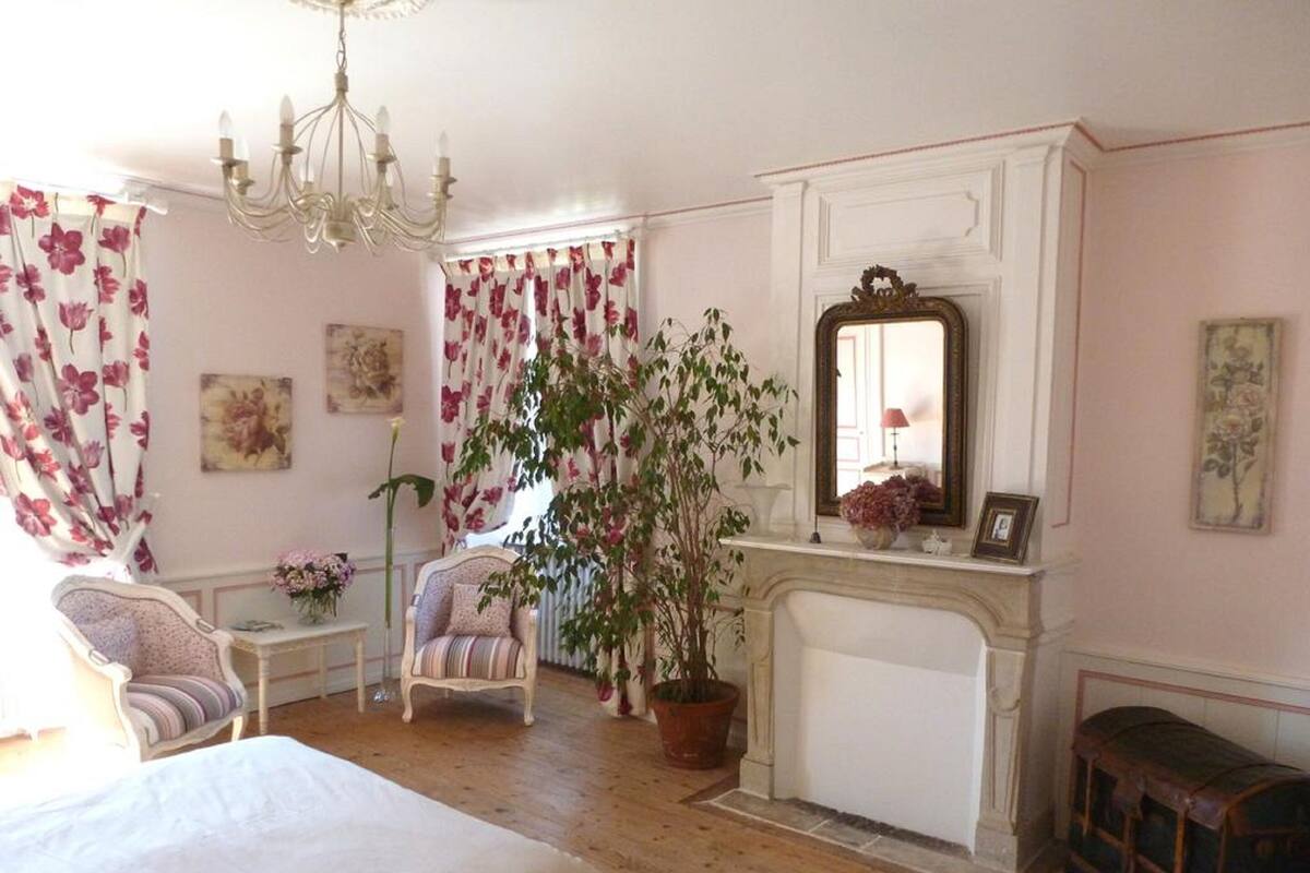 A bedroom with a bed, a chair, a table, a fireplace, and a mirror.