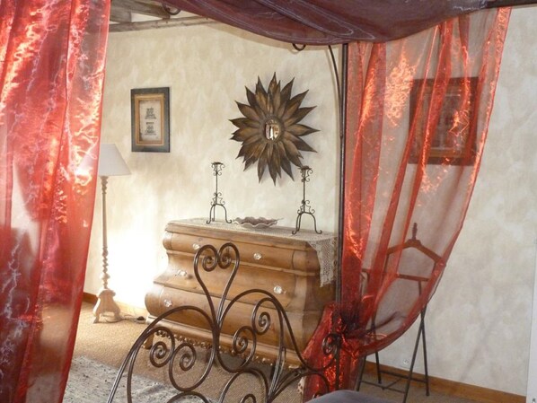 Una habitación con una cama, una mesita de noche, una lámpara, un espejo decorativo con diseño de sol y cortinas rojas.