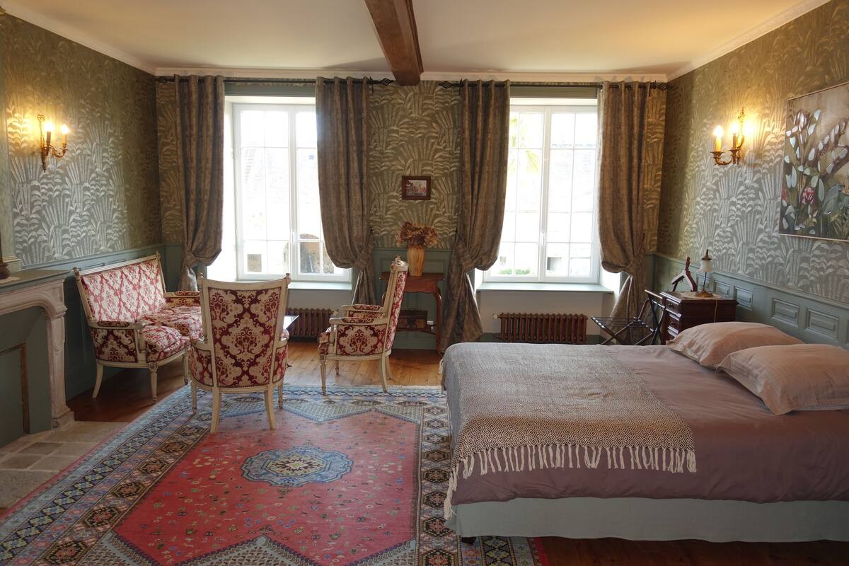 A bedroom with a large bed, two chairs, a table, and a window with curtains.