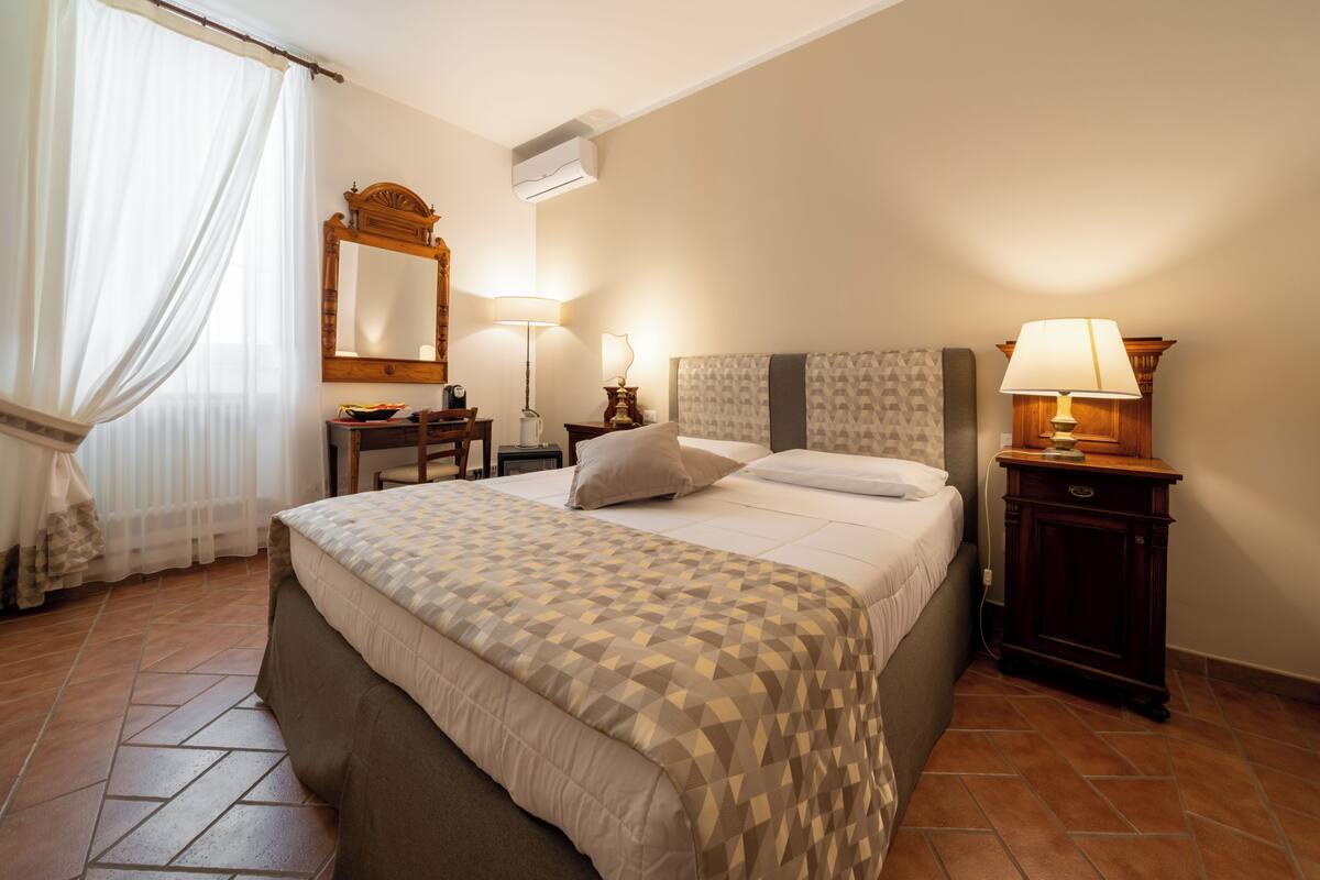 A hotel room with a bed, bedside tables, a mirror, a lamp, and a window with curtains.