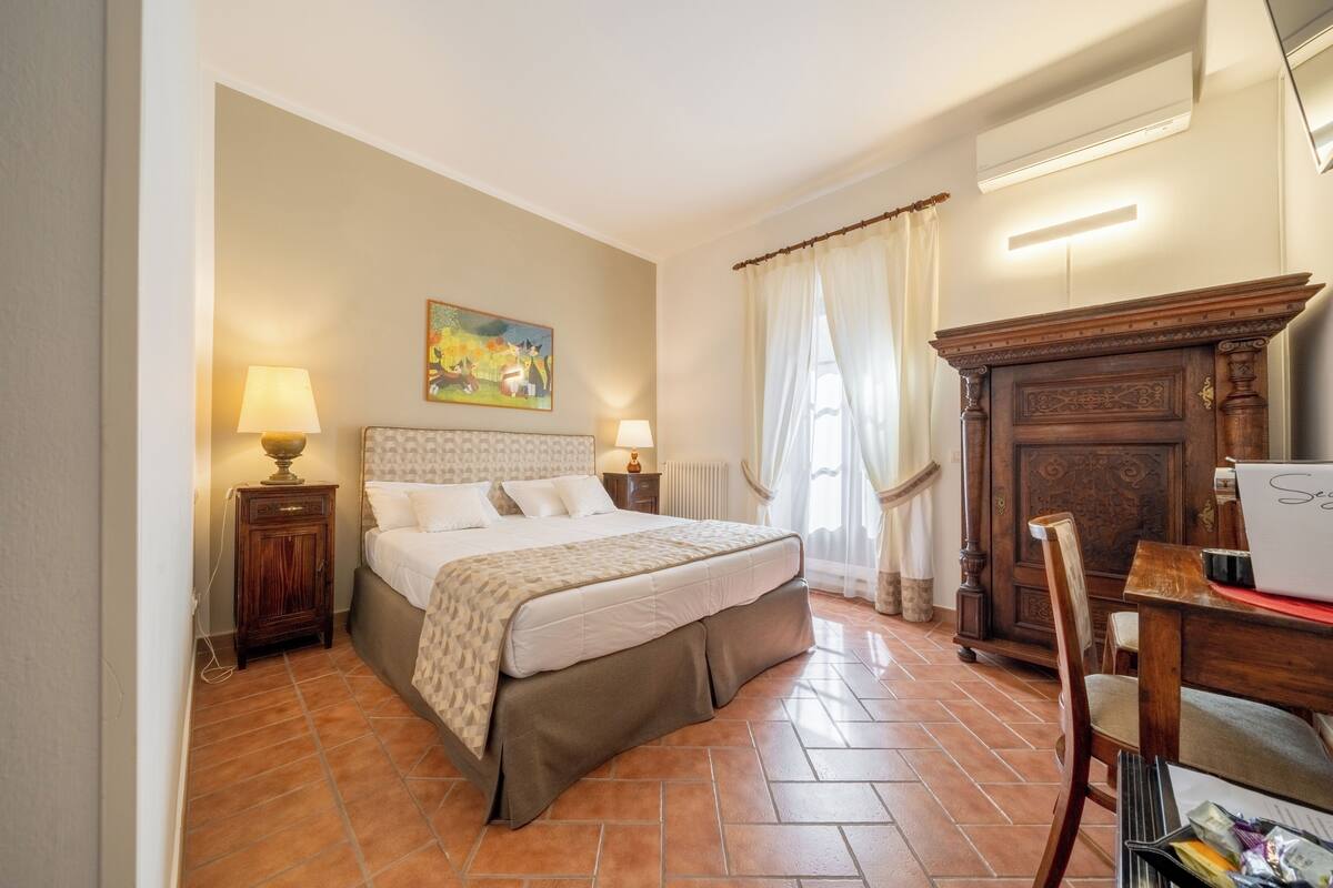 A bedroom with a bed, two bedside lamps, a painting, a wooden dresser, and a desk with a laptop.