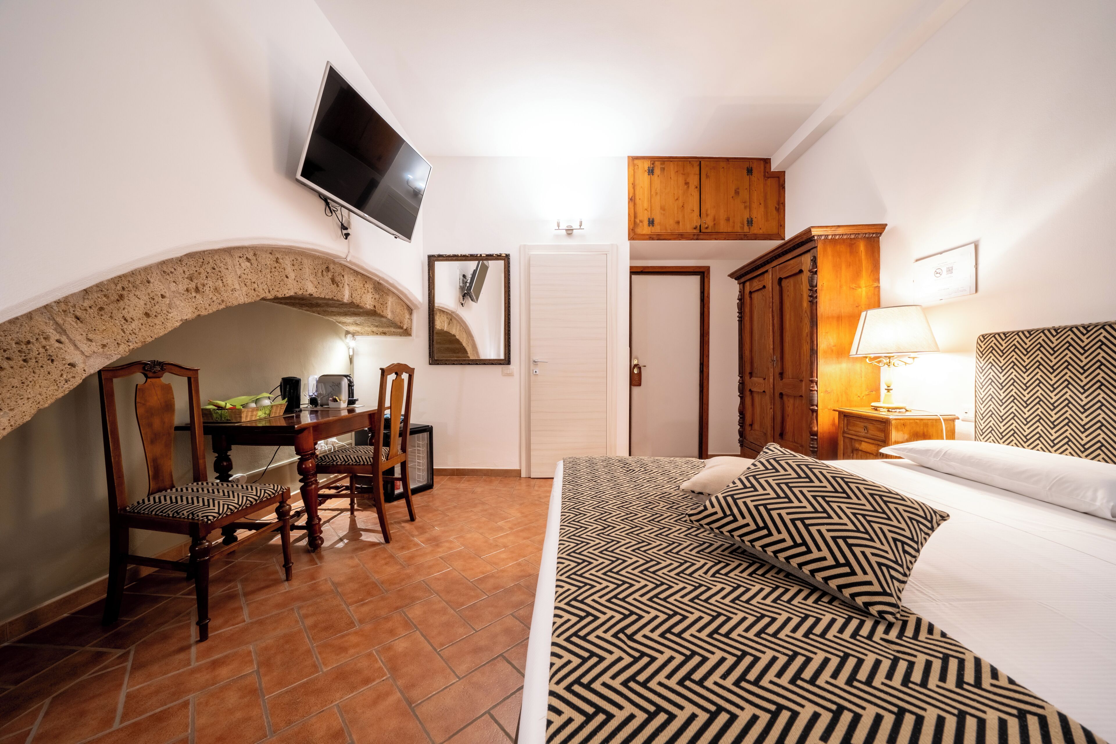 Une chambre à coucher comprenant un lit, un téléviseur fixé au mur, une armoire en bois, un bureau avec des chaises et un sol carrelé.