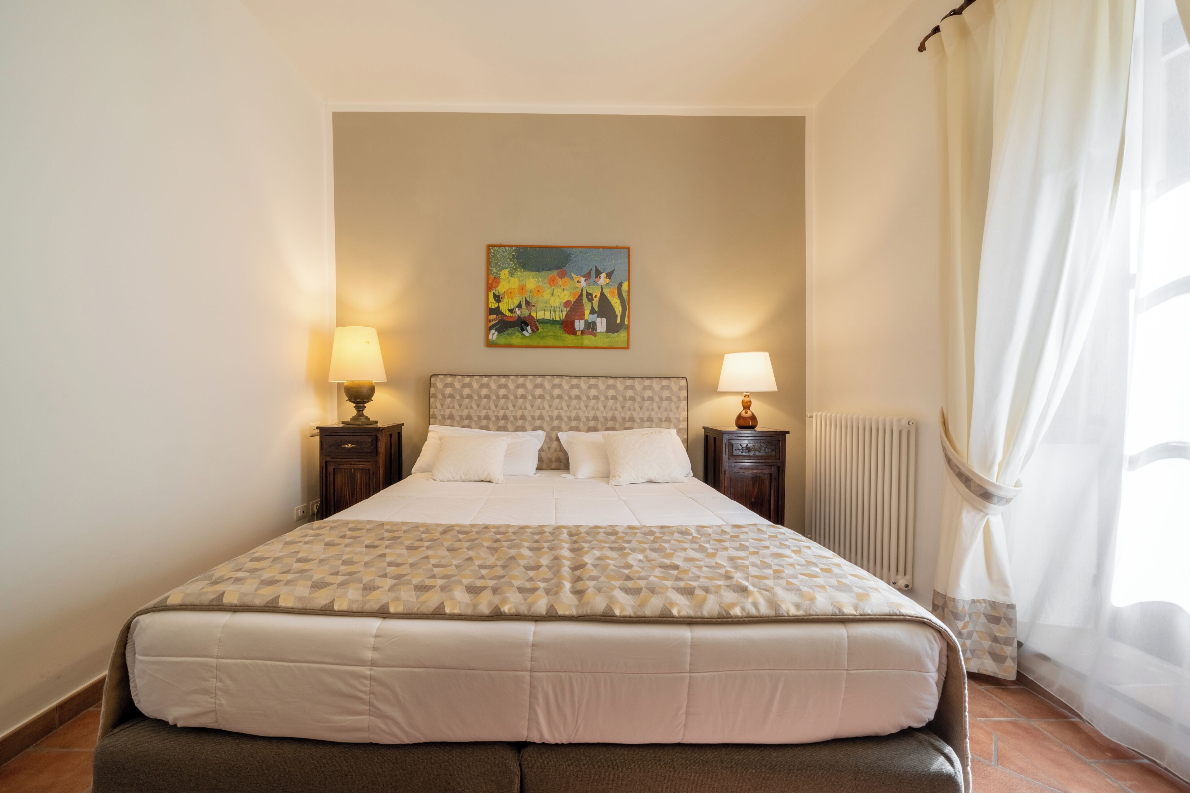 Un letto rifatto con cura, una coperta a motivi decorativi, due lampade ai lati del letto, una testiera con un motivo geometrico e un quadro appeso al muro.