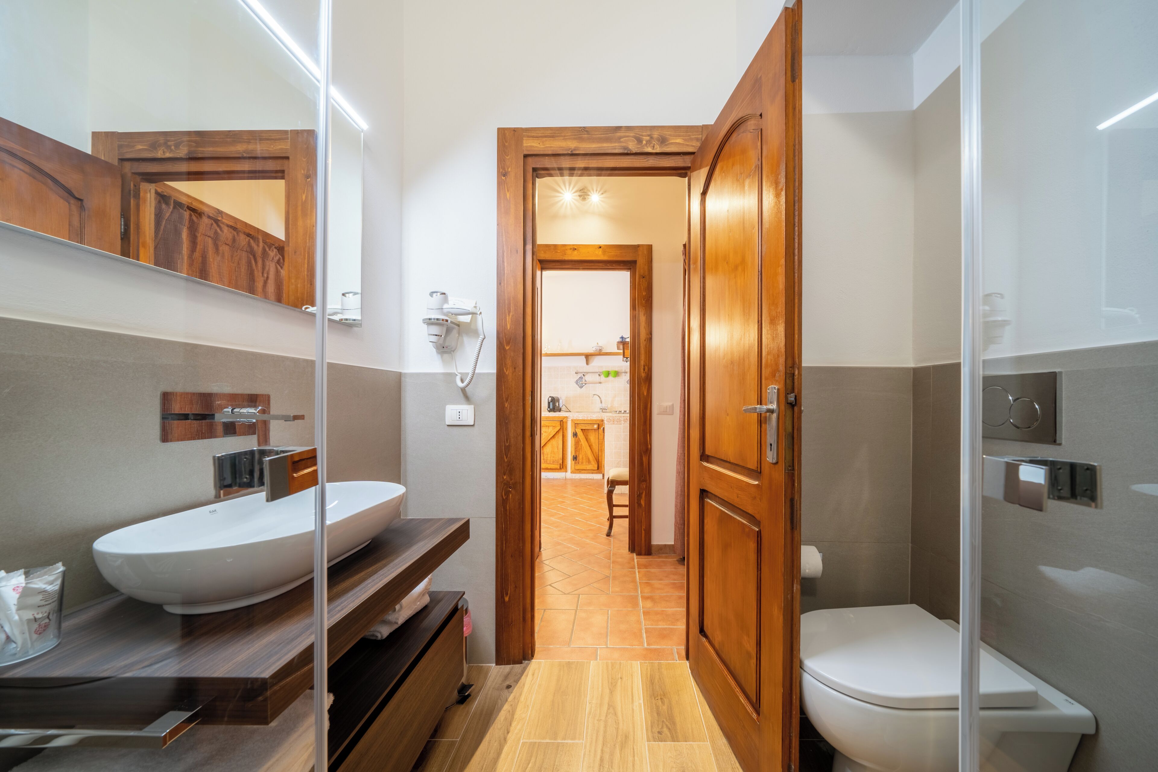 Un baño moderno con un lavabo blanco grande, una cabina de ducha de cristal y una puerta de madera que da a una zona de cocina.