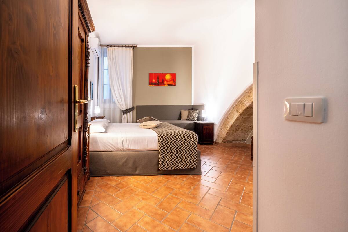 A bedroom with a bed, a nightstand, a wall-mounted light switch, and a window with curtains.