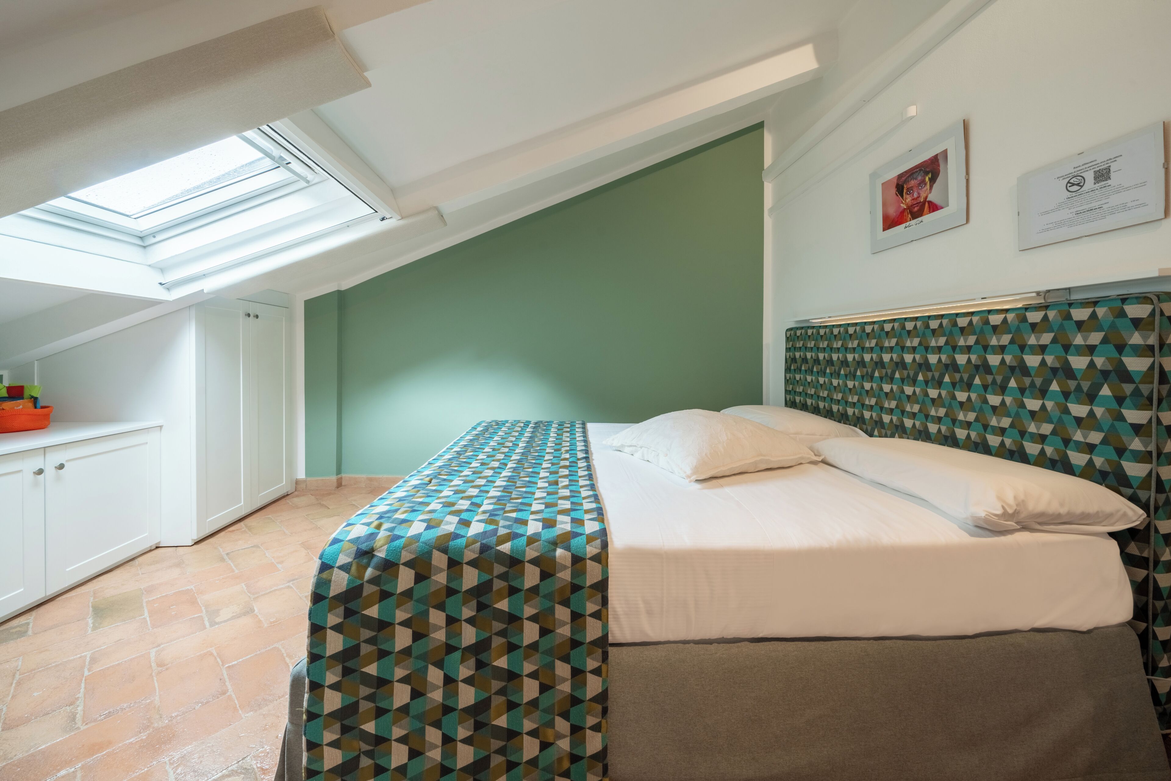 A bedroom with a skylight, a bed with patterned bedding, a green wall, and a tiled floor.