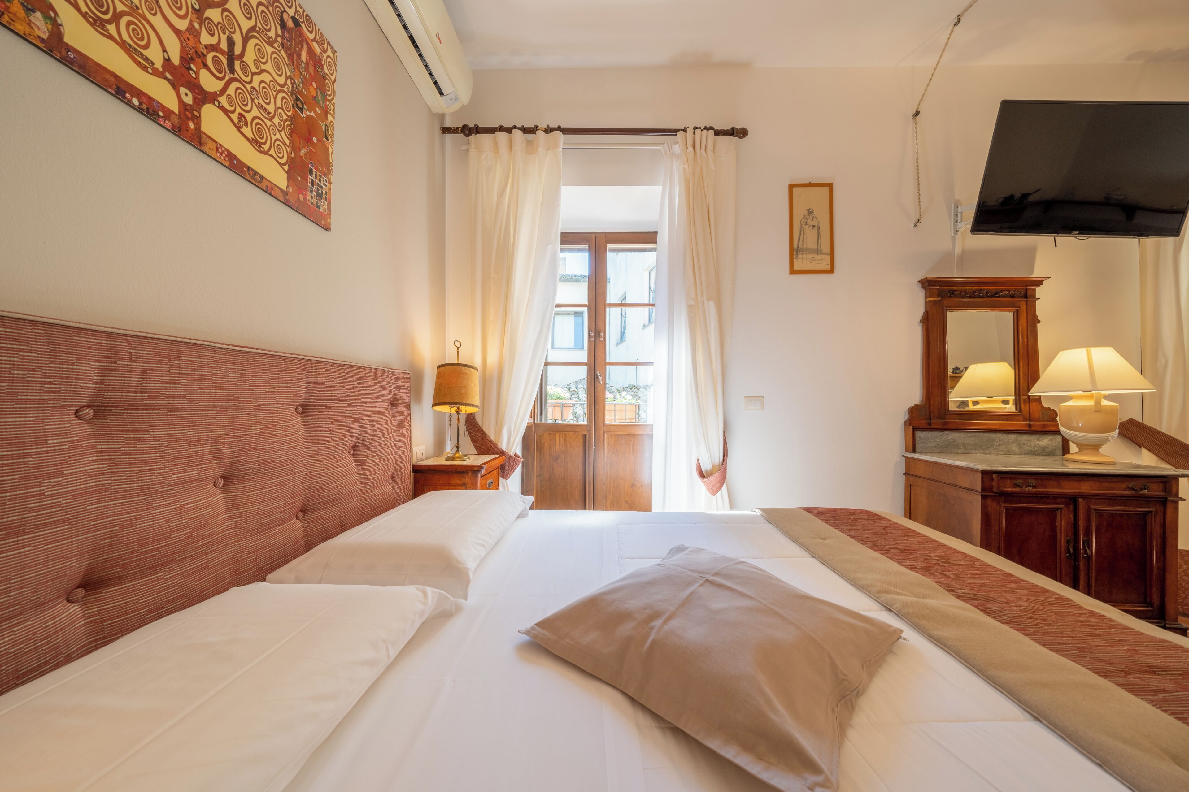 Habitación de hotel con una cama grande, cabecero de madera, televisor, escritorio con lámpara y ventana con cortinas.