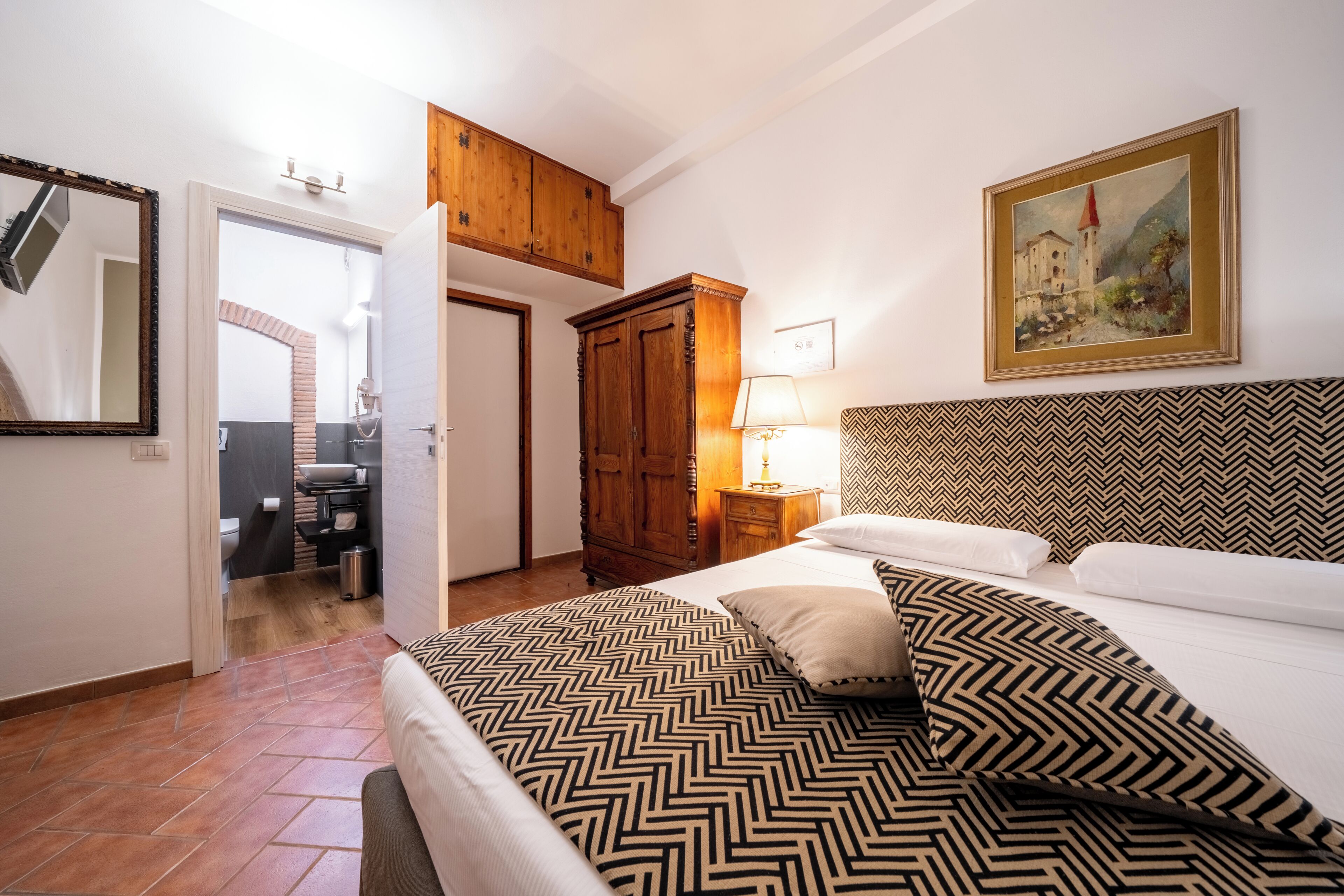 A bedroom with a bed, a wooden wardrobe, a painting on the wall, and a bathroom visible through an open door.