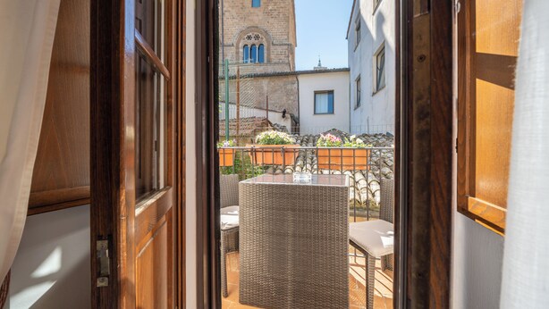 Puerta abierta que da a un balcón con muebles de mimbre y vista a una torre histórica.