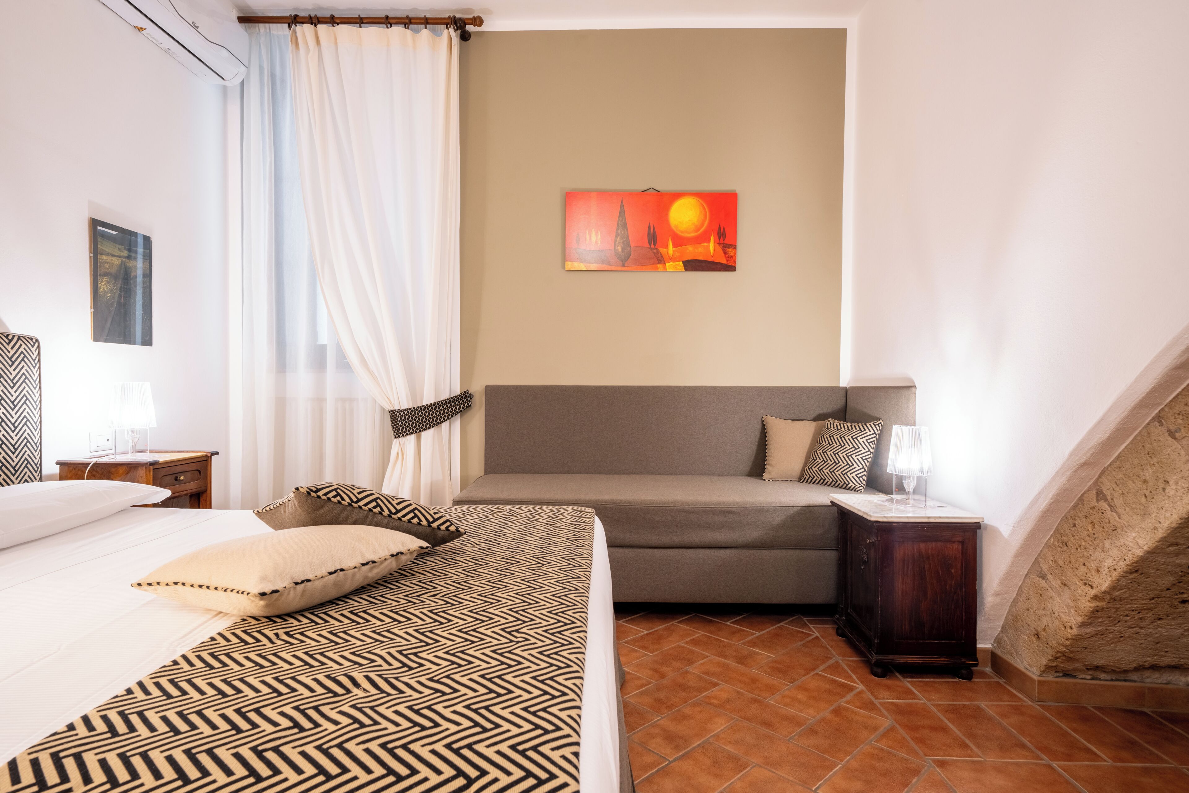 A bedroom with a bed, a sofa, a nightstand, a lamp, a painting, and a window with curtains.