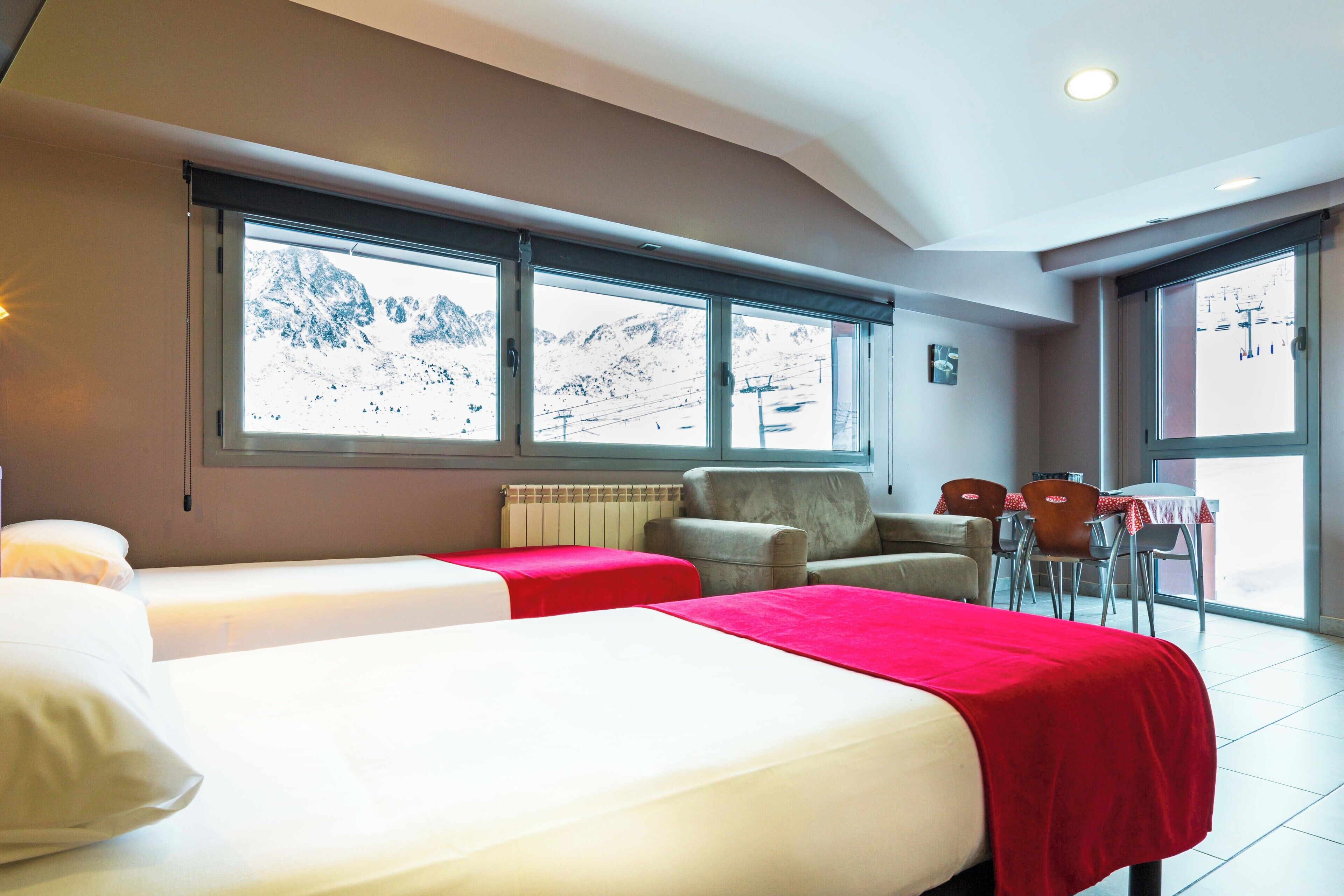 A hotel room with two beds, a red blanket, a chair, a table, and a view of mountains through large windows.