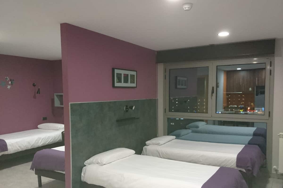 A hotel room with four beds arranged in a row, a window with a view of the city, and a small table with a chair.