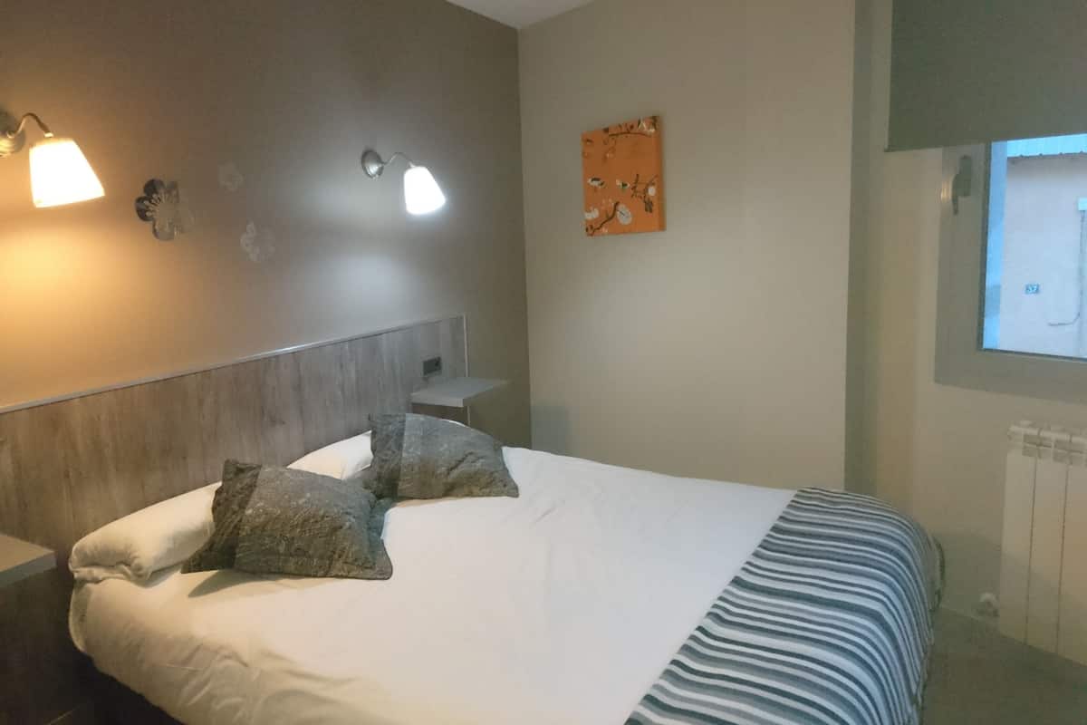 A hotel room with a bed, two pillows, a nightstand, a wall-mounted lamp, a small table, and a painting.